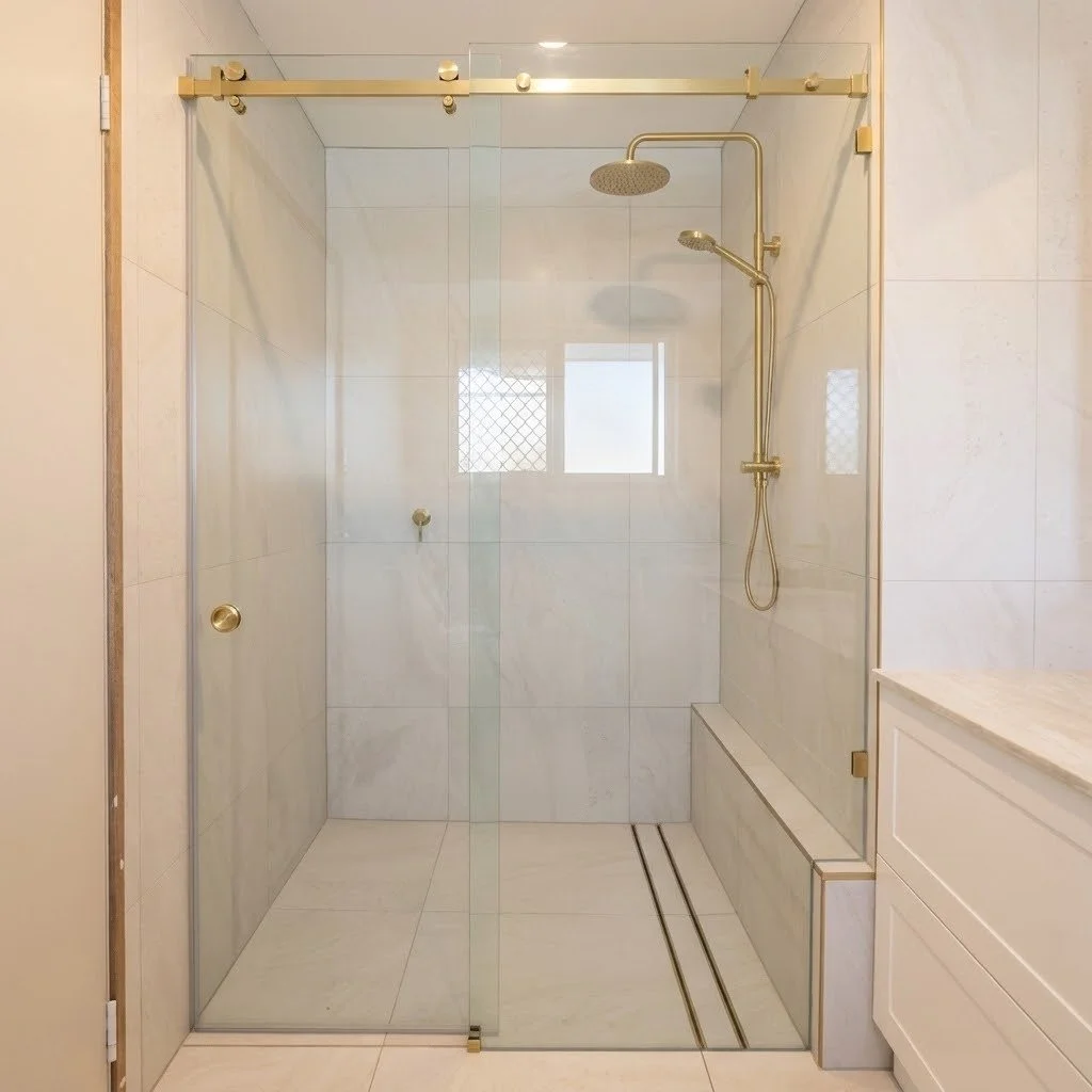 Modern walk-in shower with glass enclosure, gold fixtures, and a small window on the wall. There is a built-in bench or ledge on the right side.
