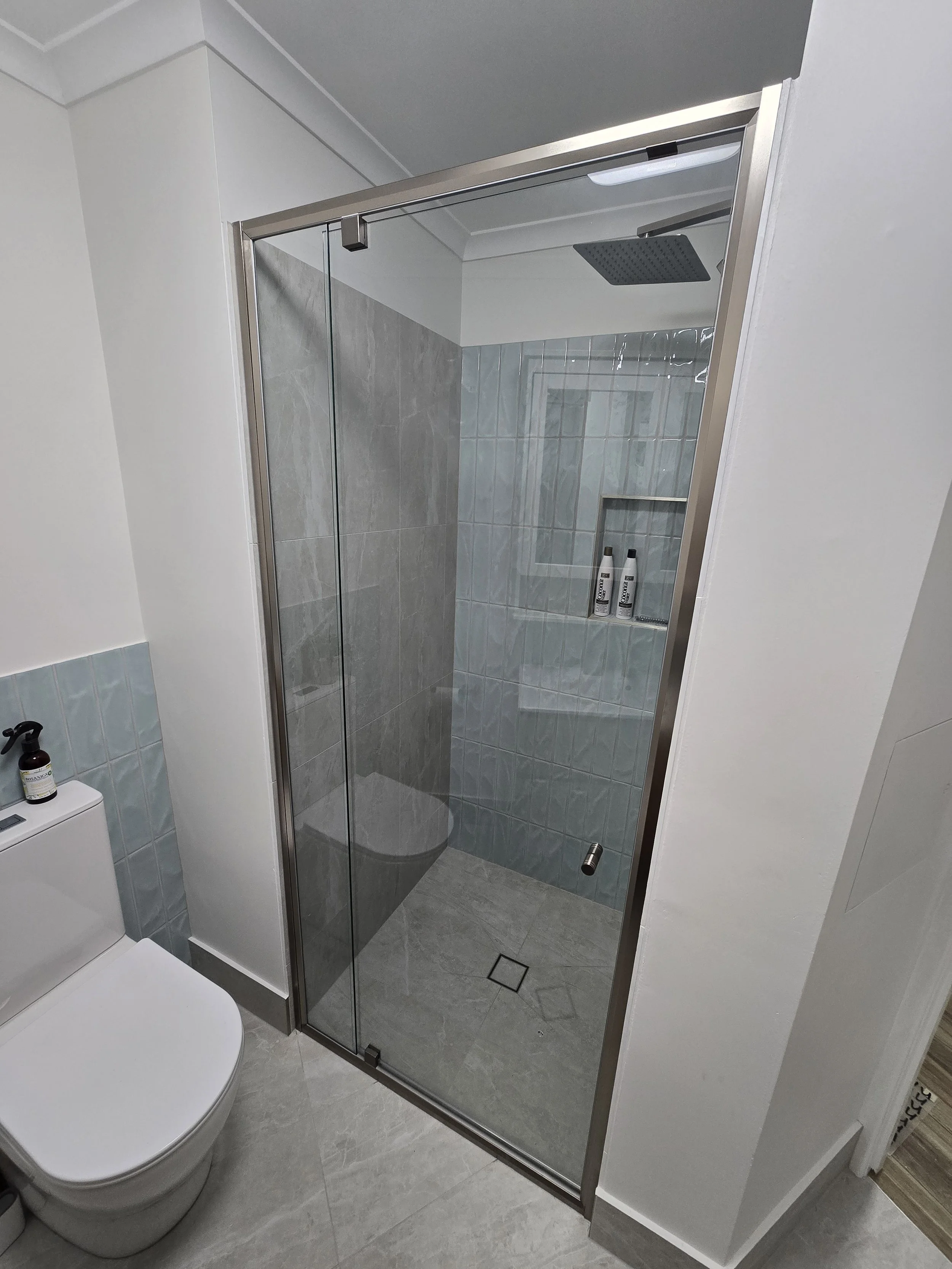 A modern bathroom with a glass shower enclosure, gray tiled walls, a white toilet, and a small shelf with two bottles inside the shower.