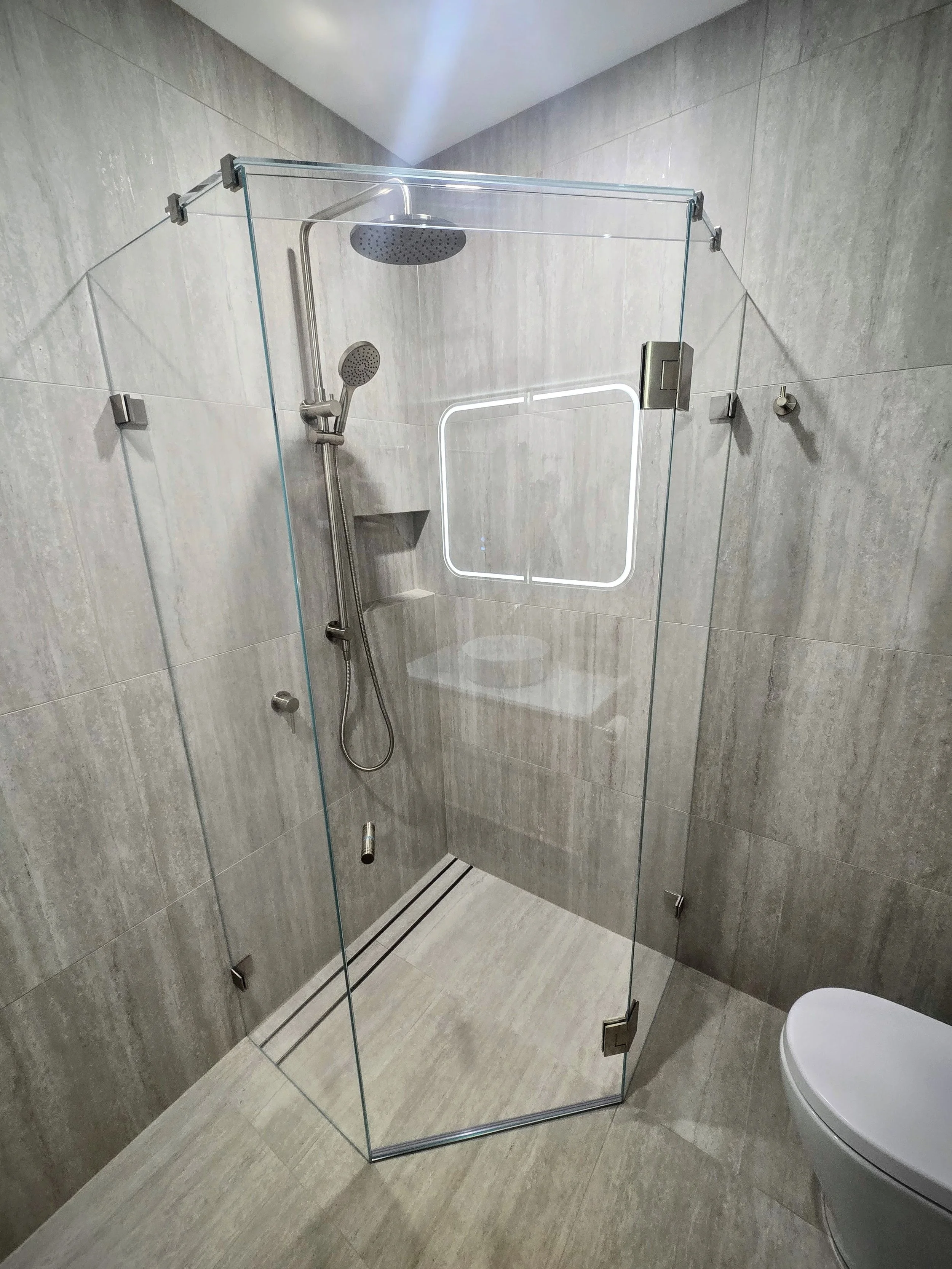 Modern bathroom shower with glass enclosure, rain shower head, handheld shower, and wall-mounted shelf, beige tiled walls and floor.