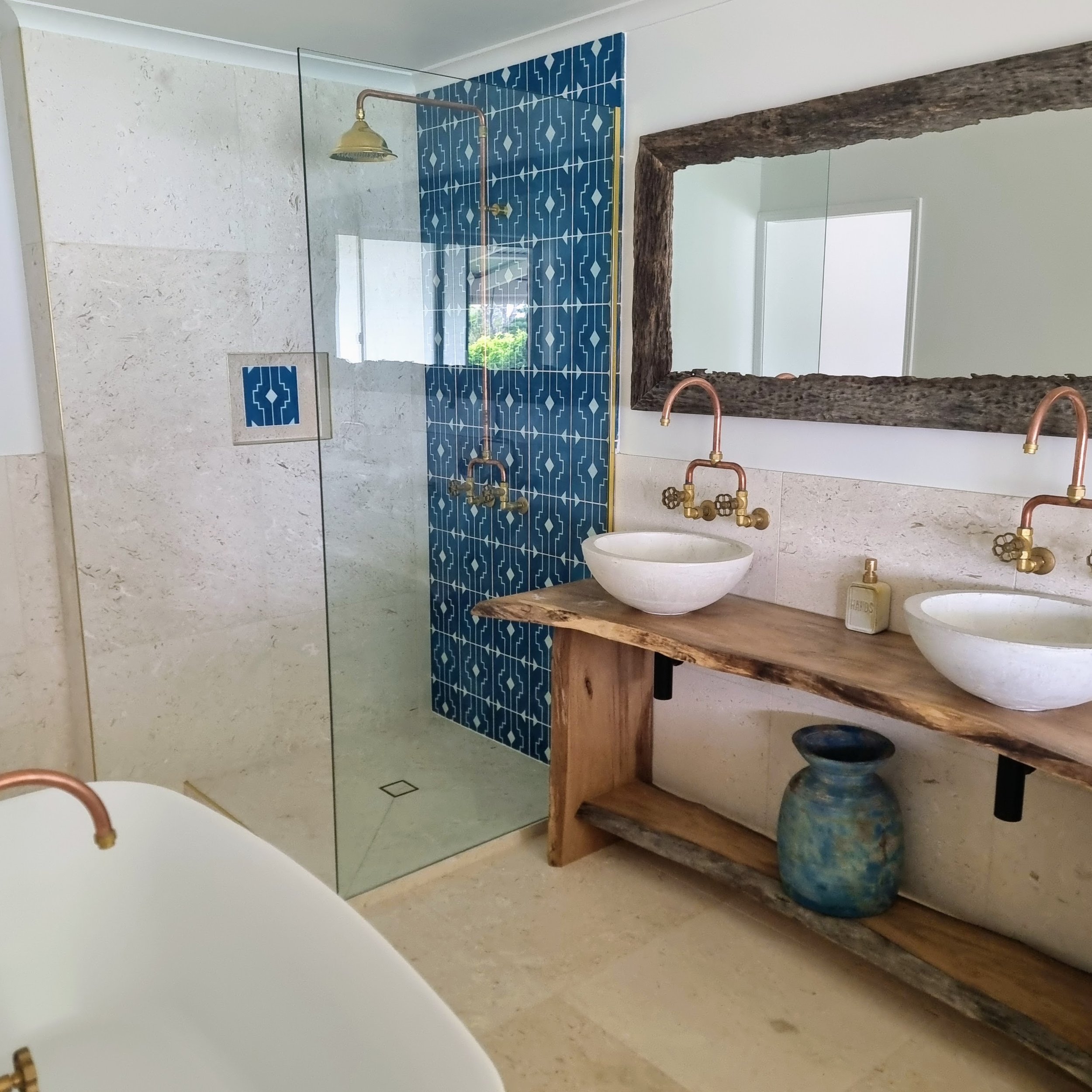 A modern bathroom featuring a walk-in shower with a glass enclosure, blue patterned tiles, a wooden vanity with two white vessel sinks, and rustic wooden mirror frames. Copper fixtures are also visible.