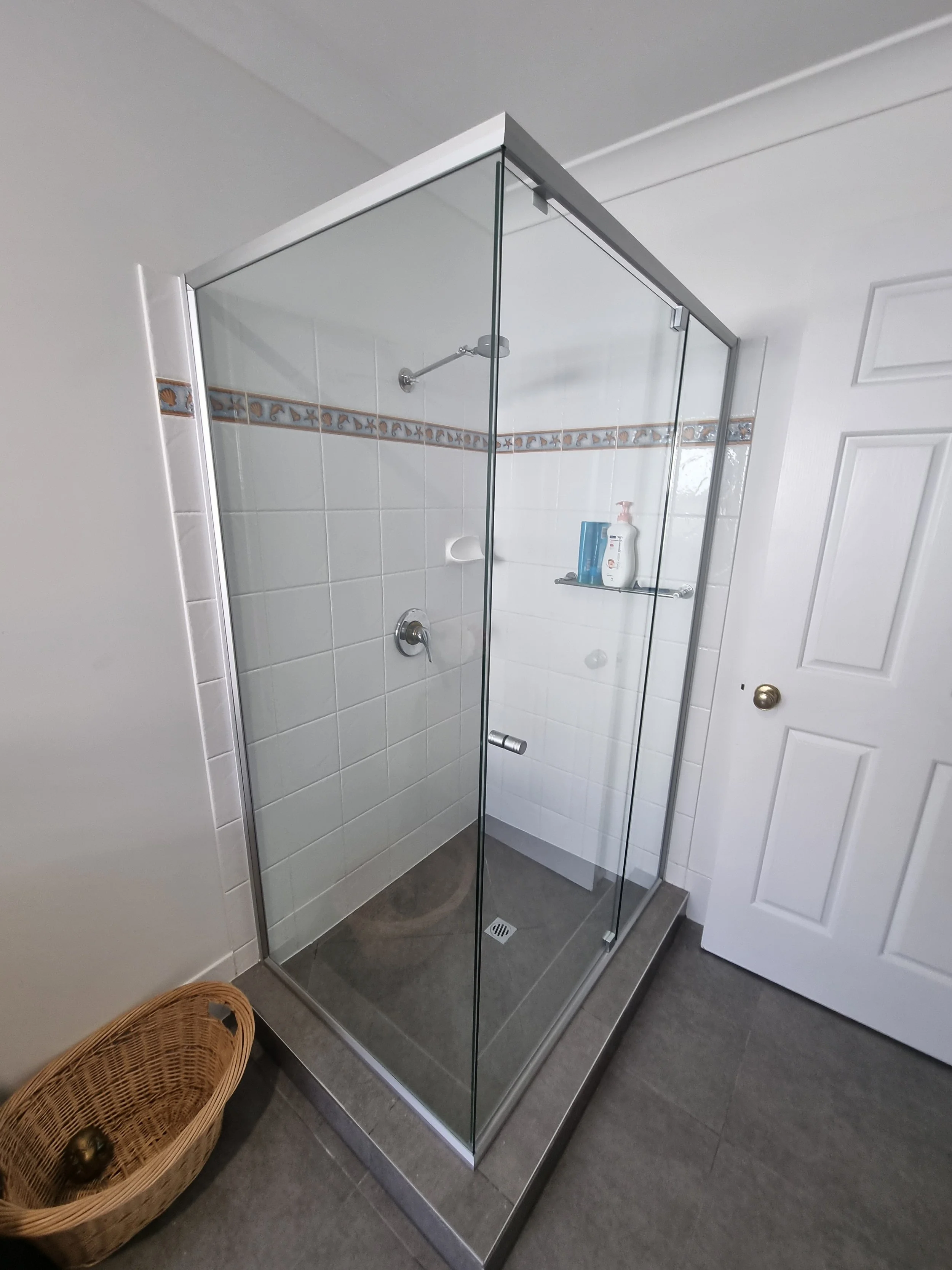 A modern corner shower enclosure with clear glass panels and a sliding glass door, framed in silver aluminium. The shower has white tiled walls with a decorative trim, a chrome shower head, a built-in soap holder, and a small glass shelf holding toil
