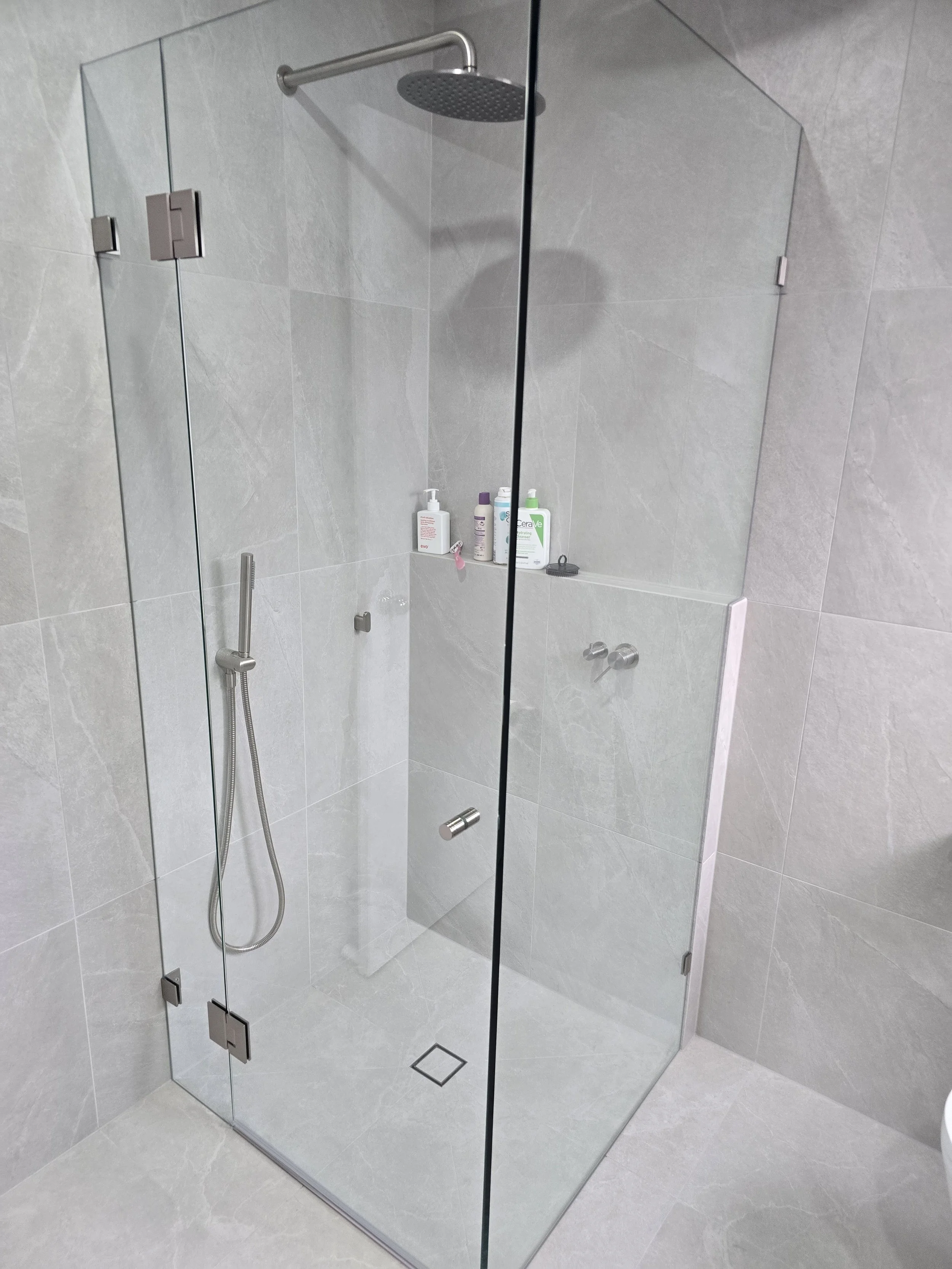 Modern glass shower enclosure with a rain shower head, grey tiled walls, and bottles of shower products on a built-in shelf.