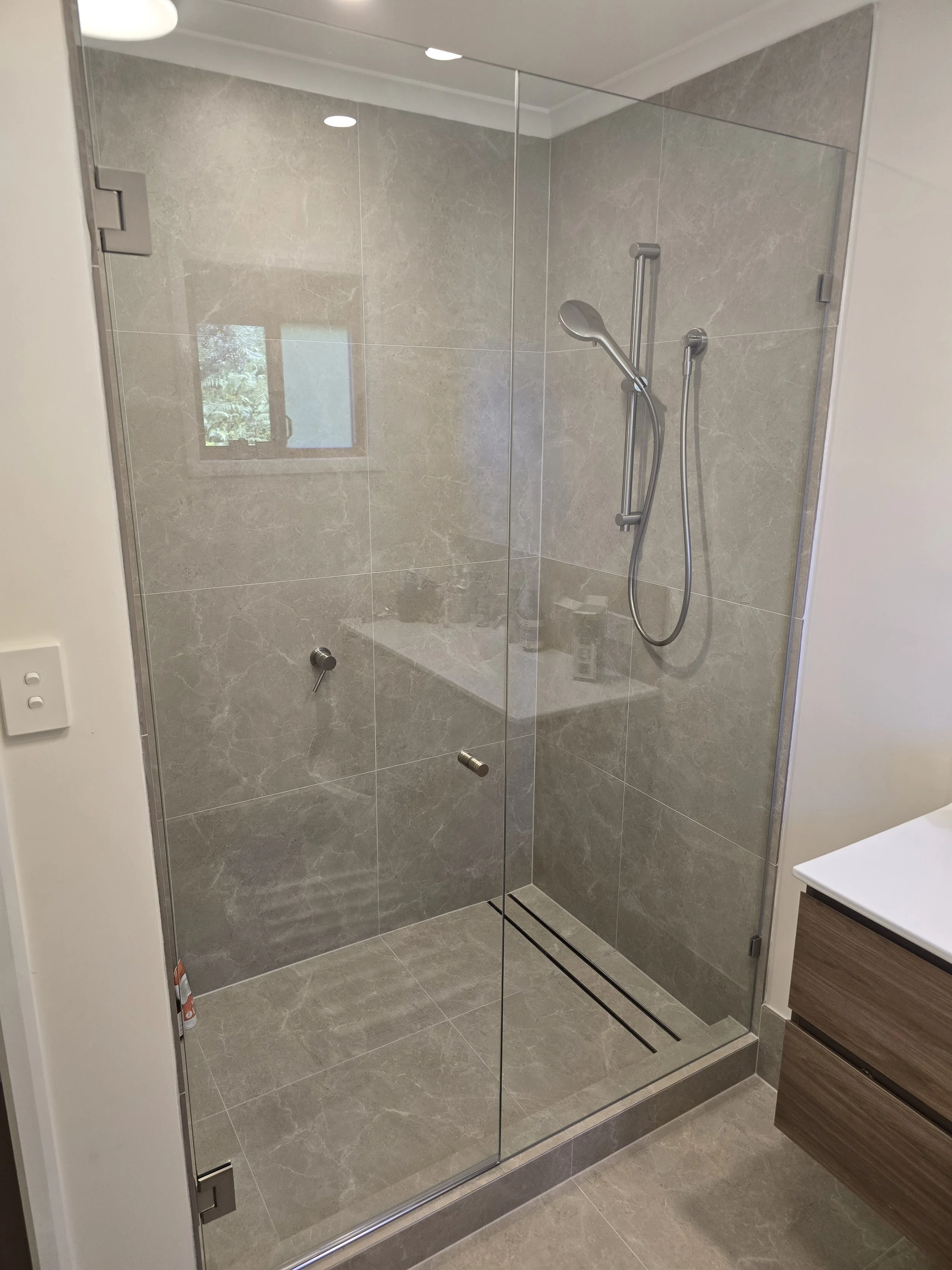 Modern walk-in shower with a glass door, gray marble tiles, a hand shower, a window reflecting trees outside, and part of a wooden bathroom cabinet.