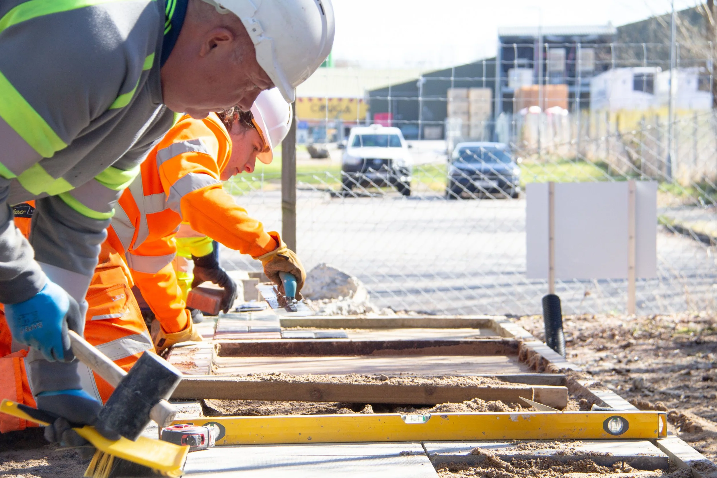 Construction workers in high-visibility clothing and helmets working on a pathway near a body of water, with a large cruise ship in the background under a cloudy sky.