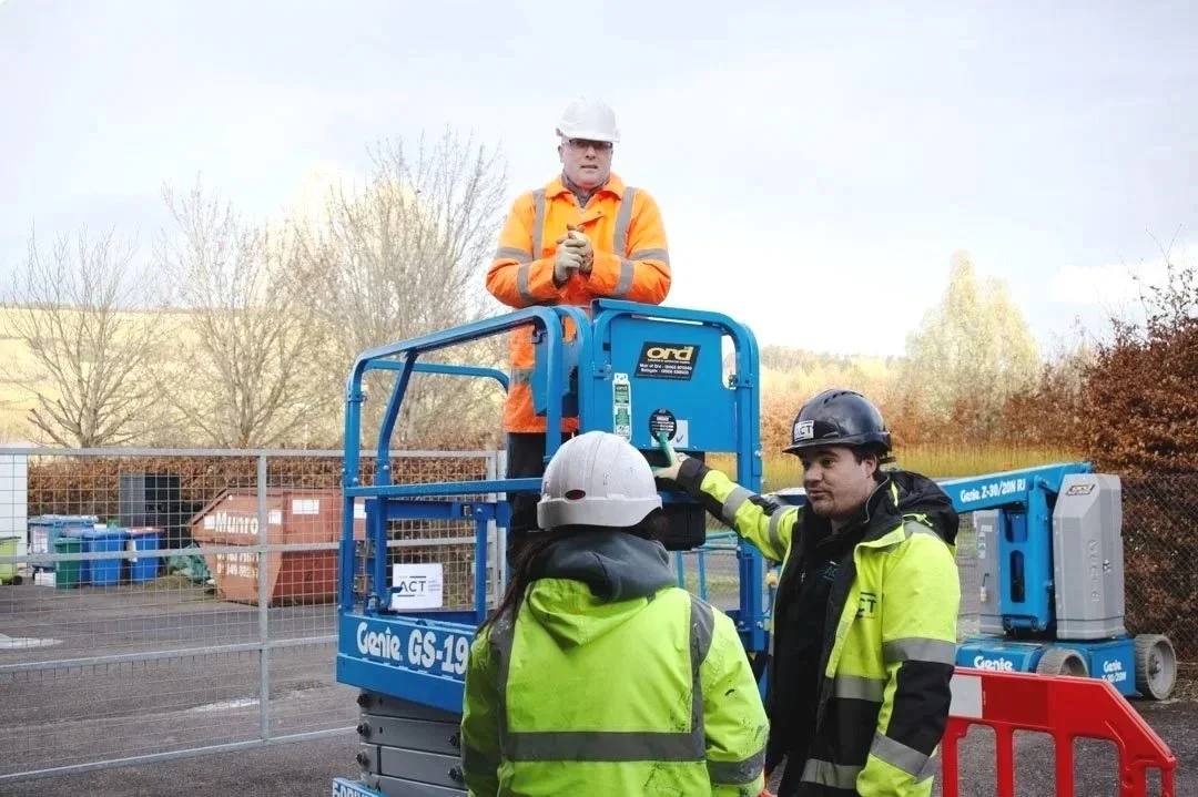 A yellow mobile aerial work platform with a scissor lift and an articulated boom lift on a sidewalk with trees and a blue sky background.