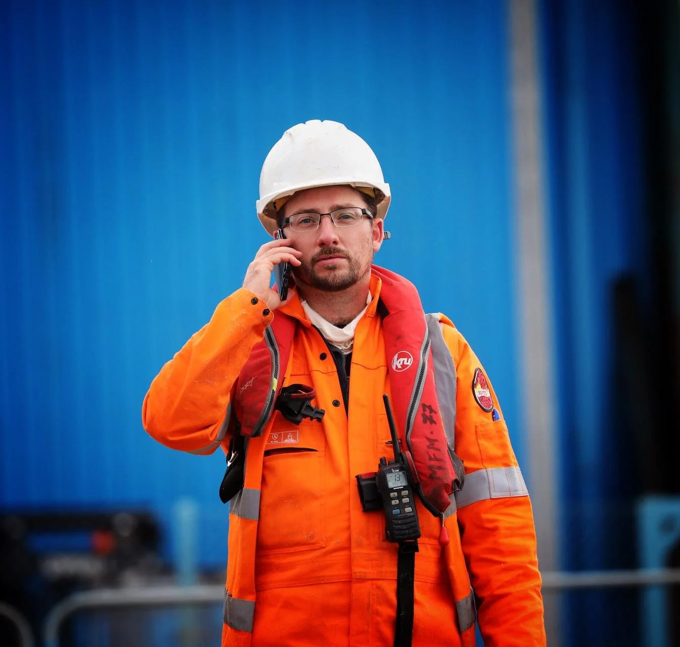 A worker in orange safety gear, wearing a white helmet, talking on a cellphone, with a blue background.