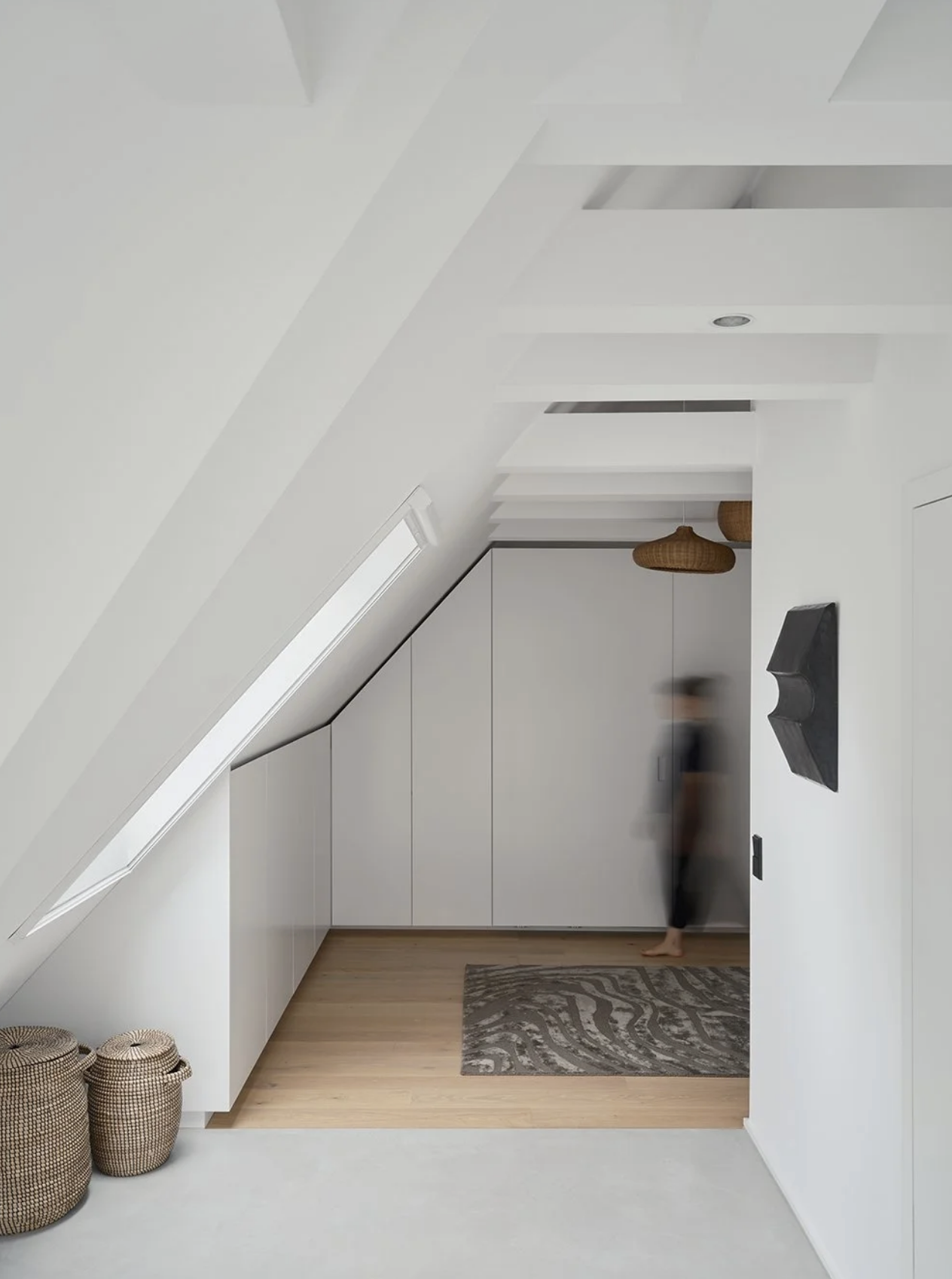 Minimalist attic room with sloped ceiling, white cabinetry, woven baskets, and a patterned rug on light wood flooring. A blurred person stands near the back.