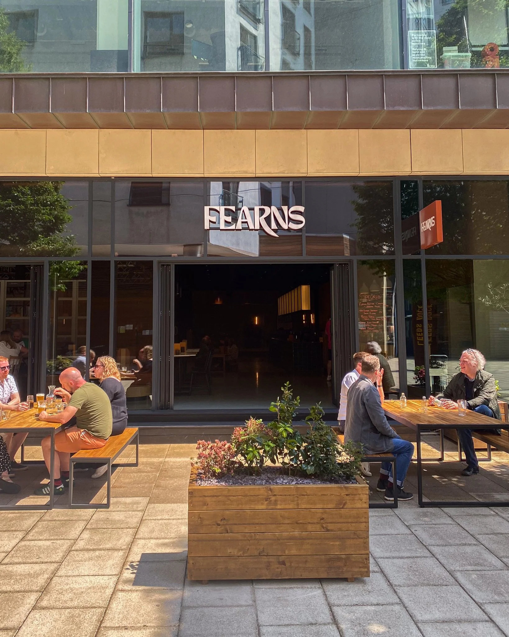 Outdoor patio of a restaurant or café with people sitting at tables and talking. A large window with the restaurant's name, 'FEARNS,' is visible on the building behind them. There are plants and trees around, and sunlight is shining on the scene.