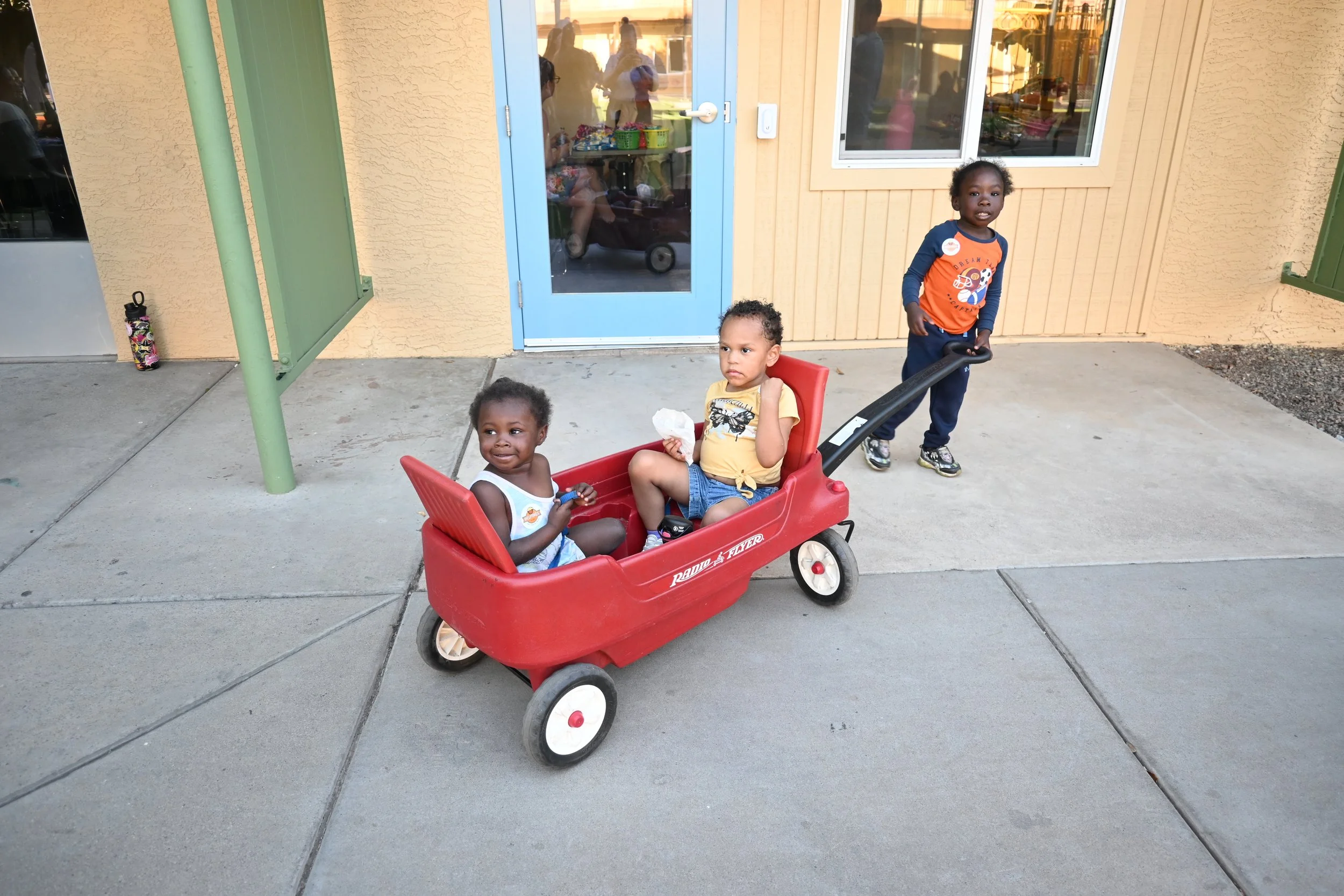 Two children sit in a red Radio Flyer wagon being pulled by another child at the UMOM courtyard