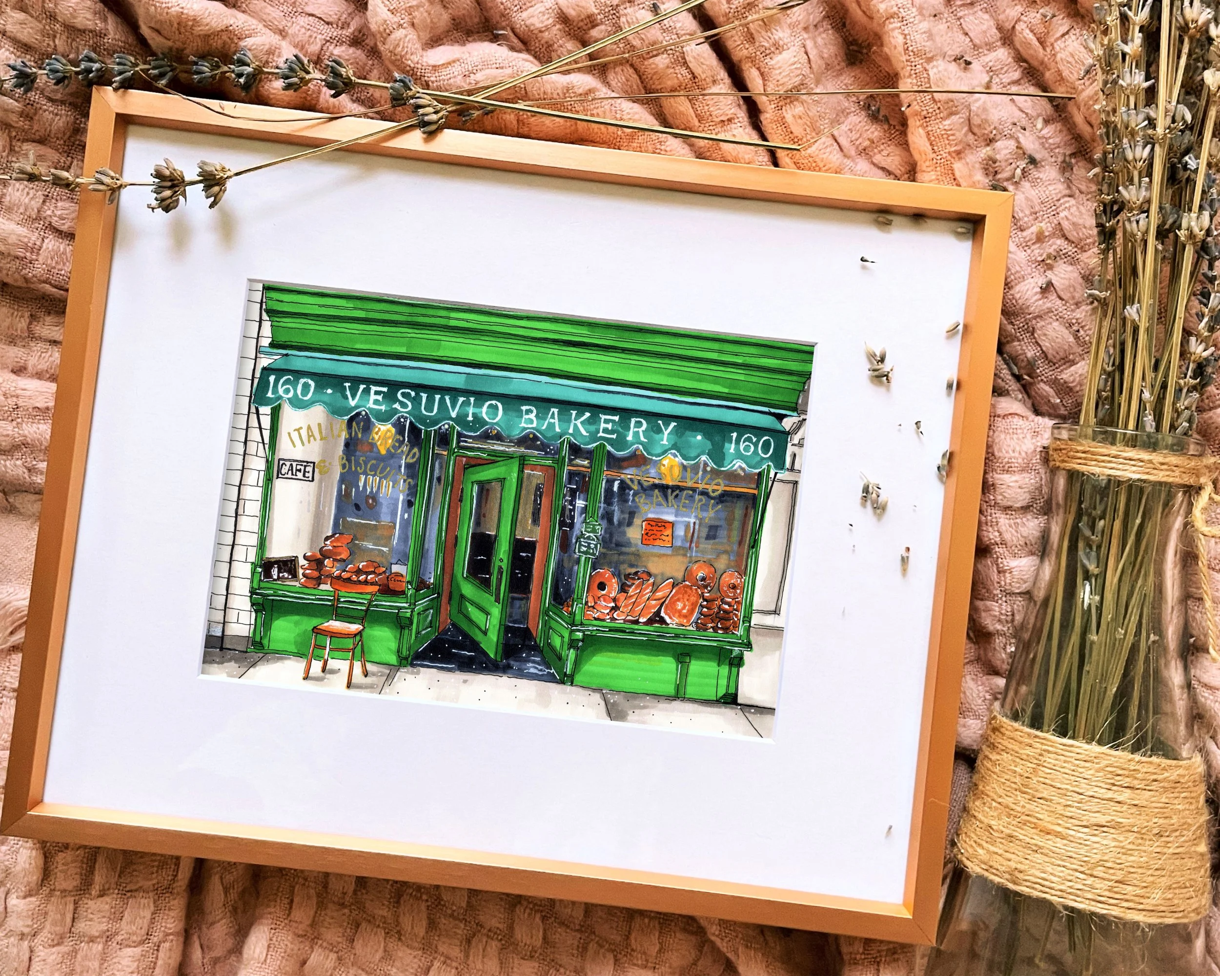 Framed in gold, this hand-illustrated drawing of Vesuvio Bakery print sits on a pink blanket