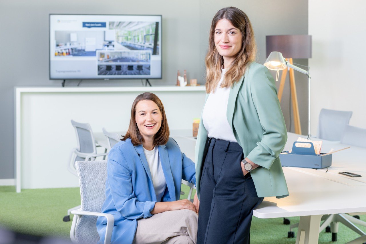 Zwei Frauen werden in einem Büro gezeigt. Sie tragen Jackets in Pastellfarben.