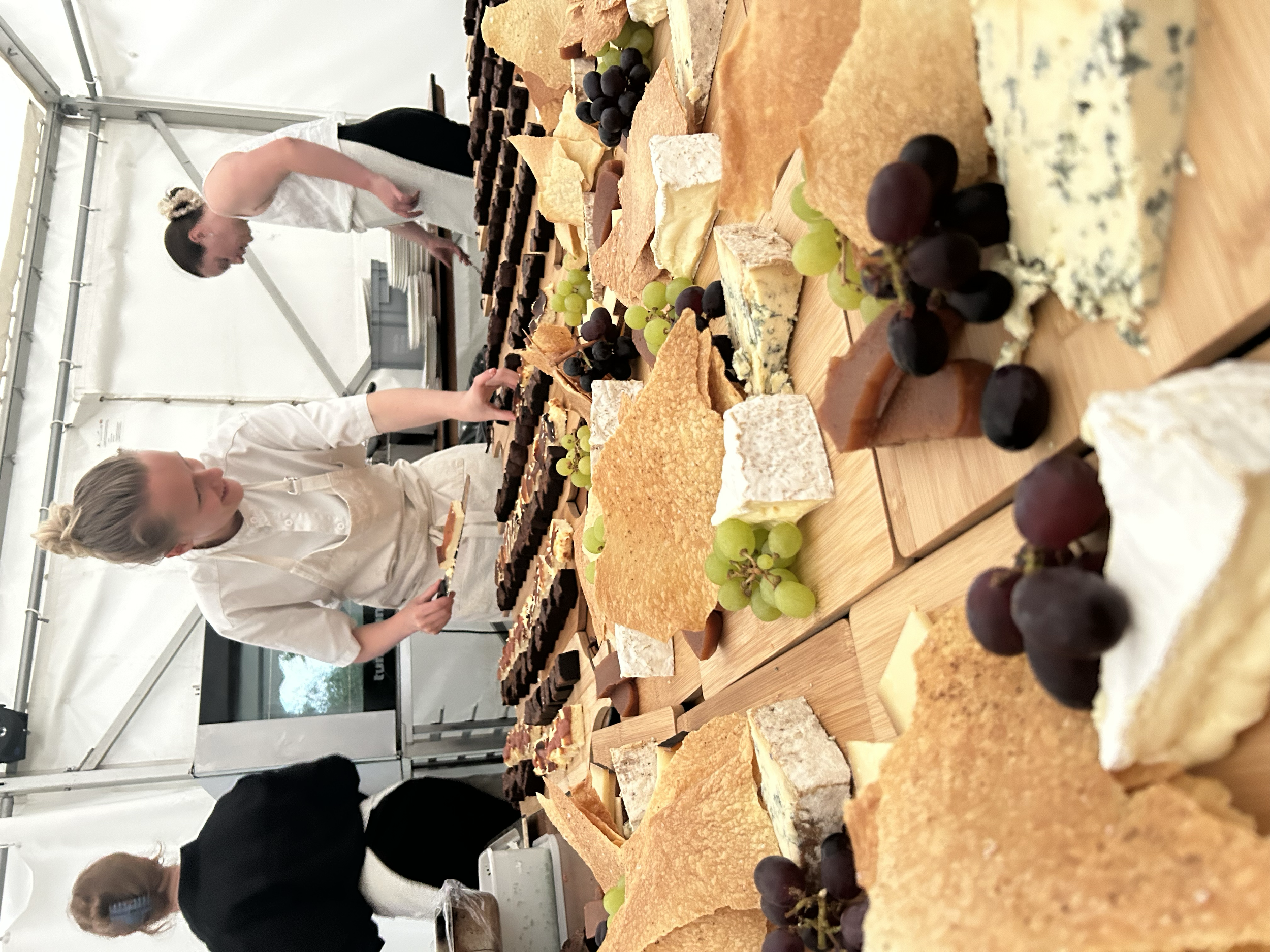 People browsing a large selection of cheeses and chocolates on a table at an indoor market or event, with some grapes and other snacks.
