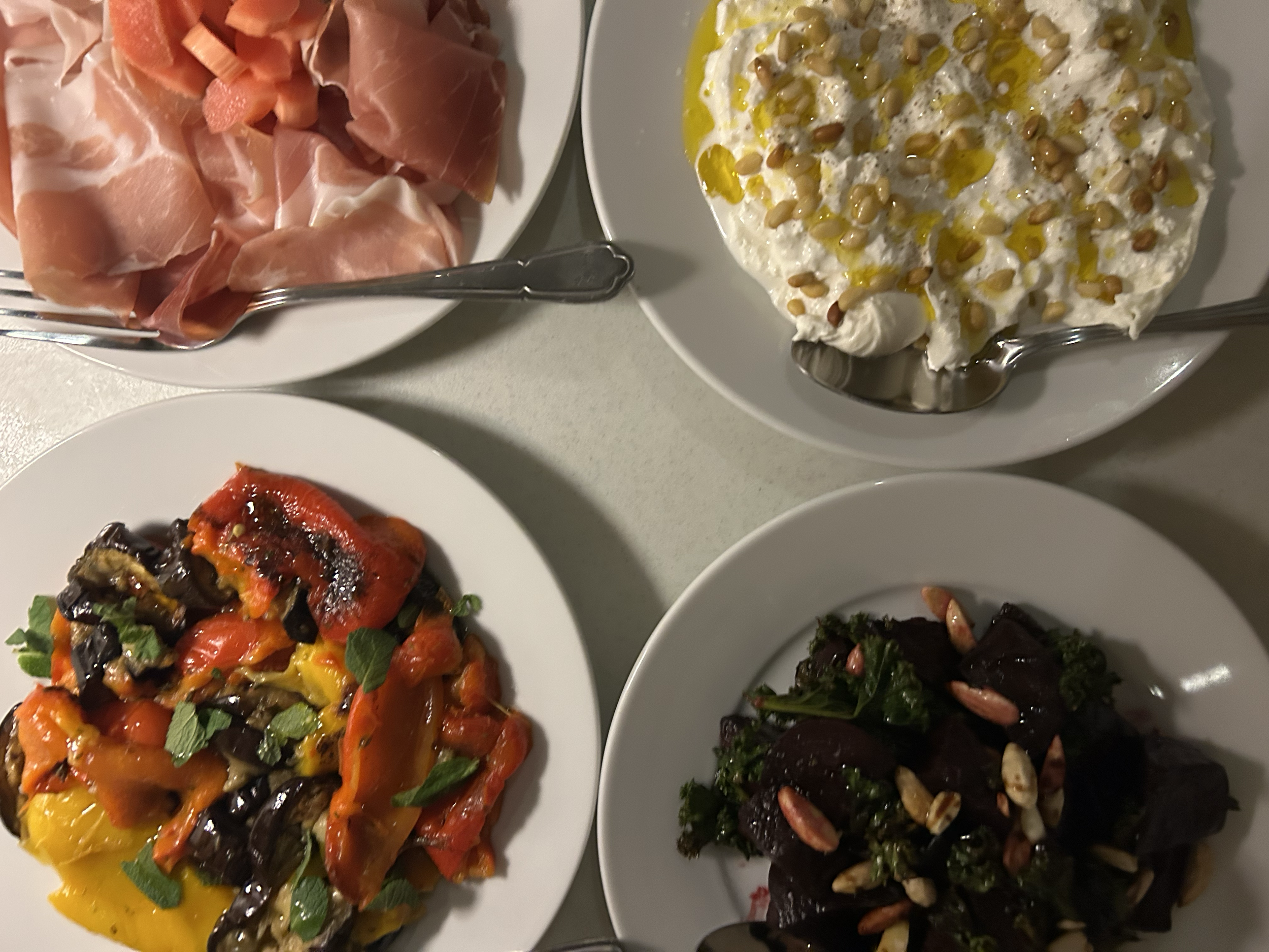 A white plate with different types of roasted or grilled vegetables, including red bell peppers, yellow bell peppers, and eggplant, garnished with green herbs. A side dish of prosciutto with parchment paper and a pair of tongs is visible on the table