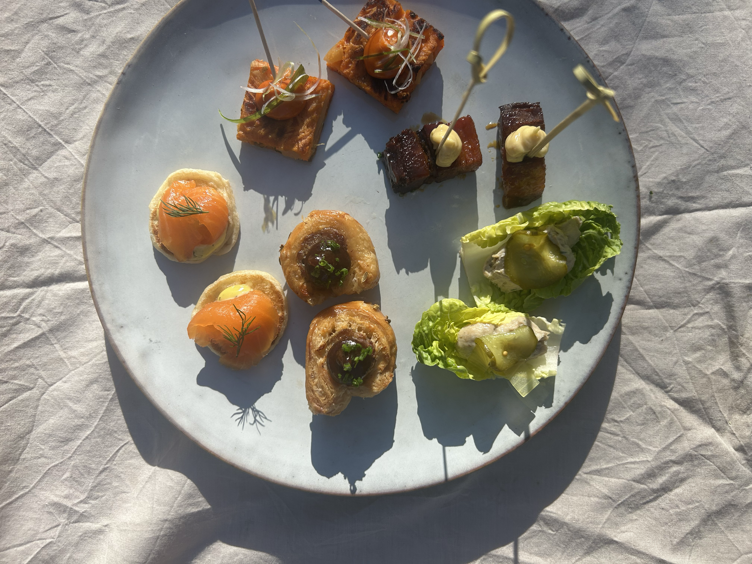 A white round plate with various small appetizers, including smoked salmon on toast, skewered meat or vegetable bites, small salad with lettuce and cucumber, and other bite-sized hors d'oeuvres, set on a textured tablecloth.