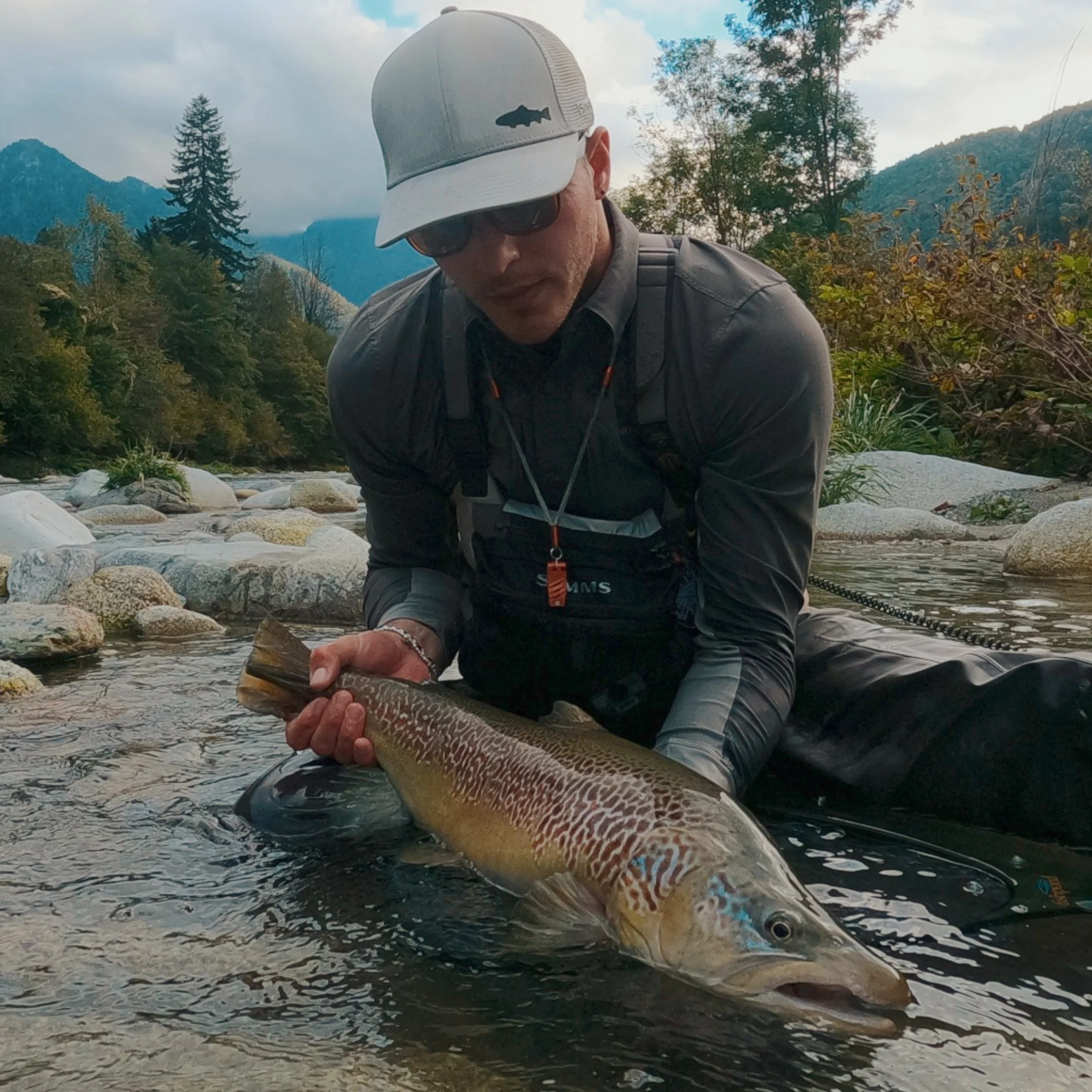 Stefano Fedrizzi wearing a gray and white cap, sunglasses, and outdoor clothing is holding a large fish in a shallow river with rocks, trees, and mountains in the background.