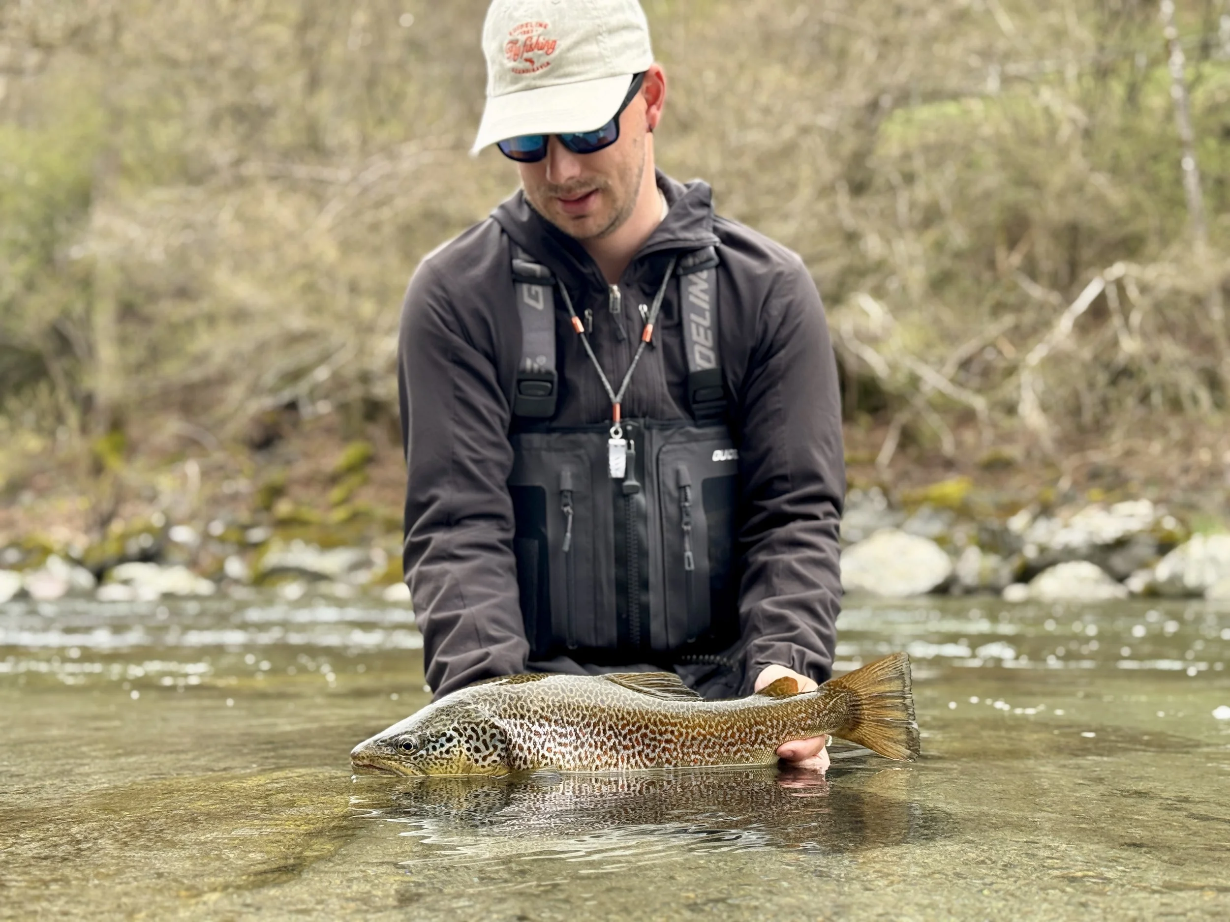 Man wearing a cap, sunglasses, and outdoor gear holding a large fish in a shallow stream.