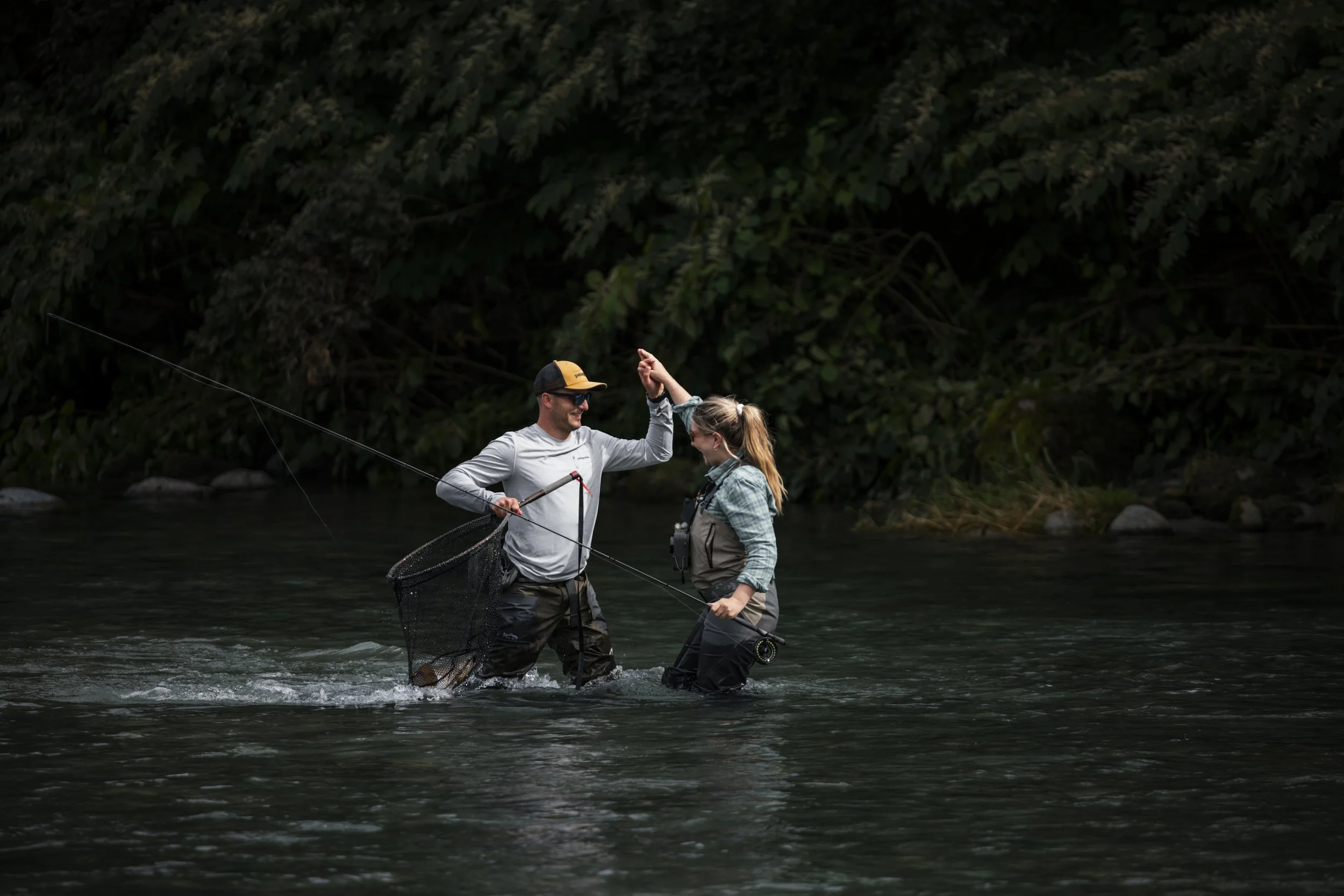 A man and woman stand in a river, fishing, with the woman smiling and holding a fishing rod while the man high-fives her, and both are dressed in outdoor fishing gear.