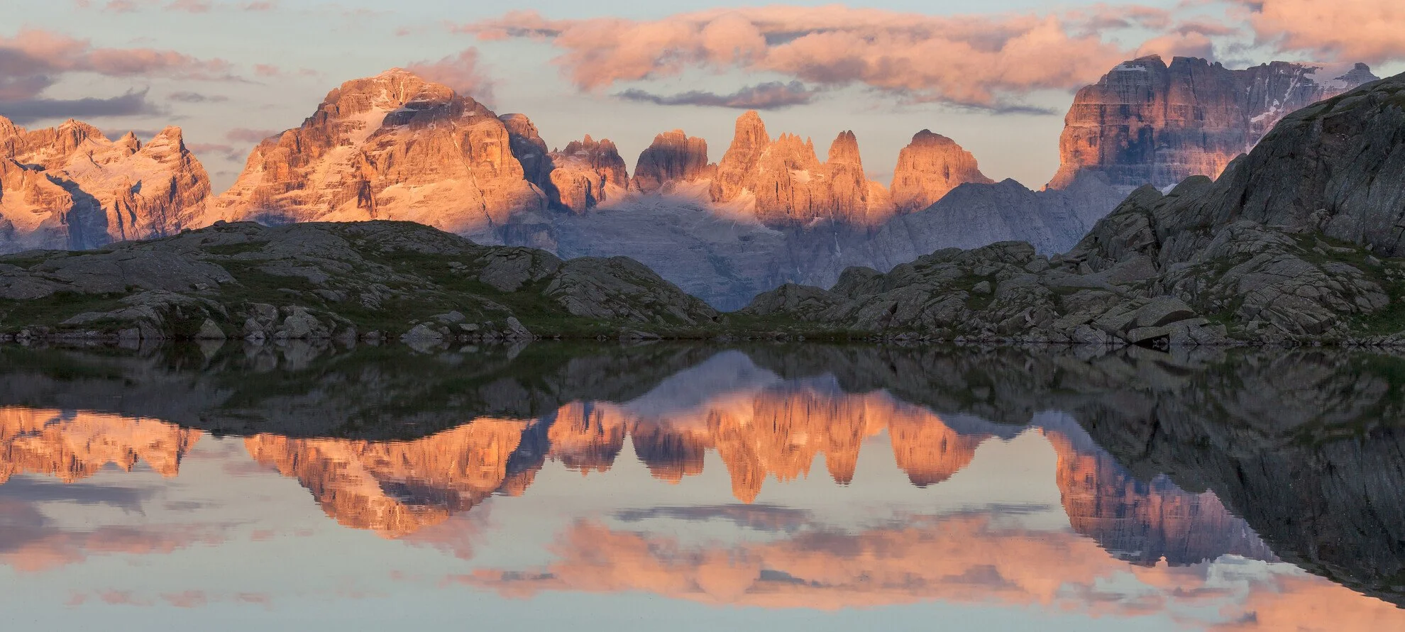Sunset over rugged Brenta dolomites with reflection in calm lake, clouds in sky