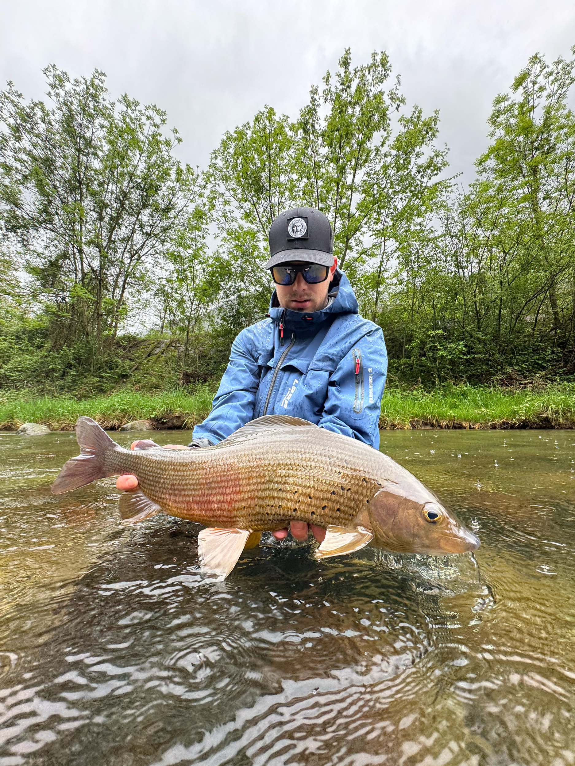 A man wearing sunglasses, a blue jacket, and a black cap holding a fish in a shallow river with trees in the background.