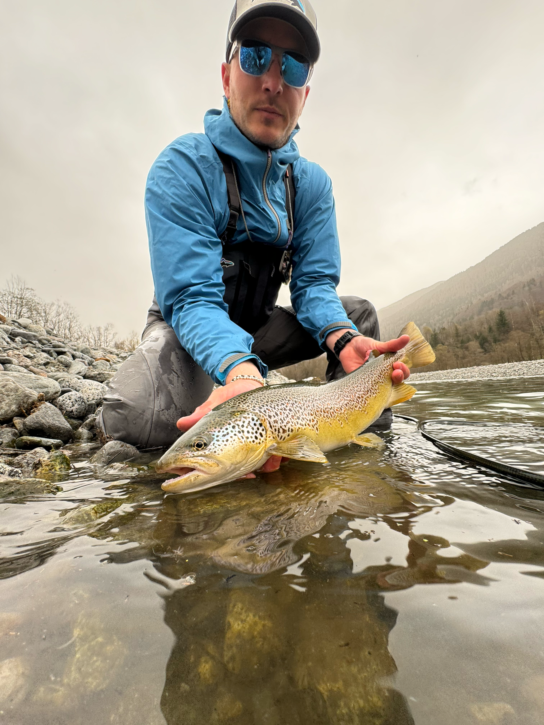 A man in outdoor gear, wearing sunglasses and a cap, kneeling by a riverbank holding a large rainbow trout.