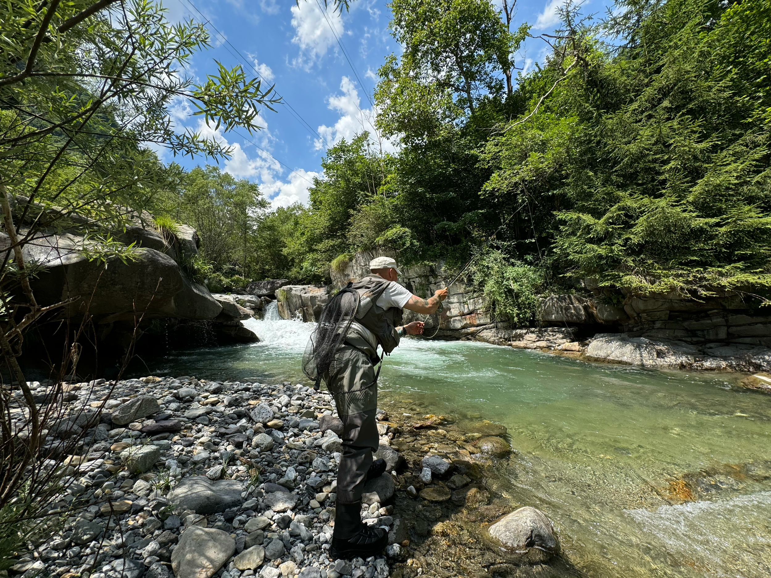 A person fishing in a river surrounded by lush green trees on a sunny day.