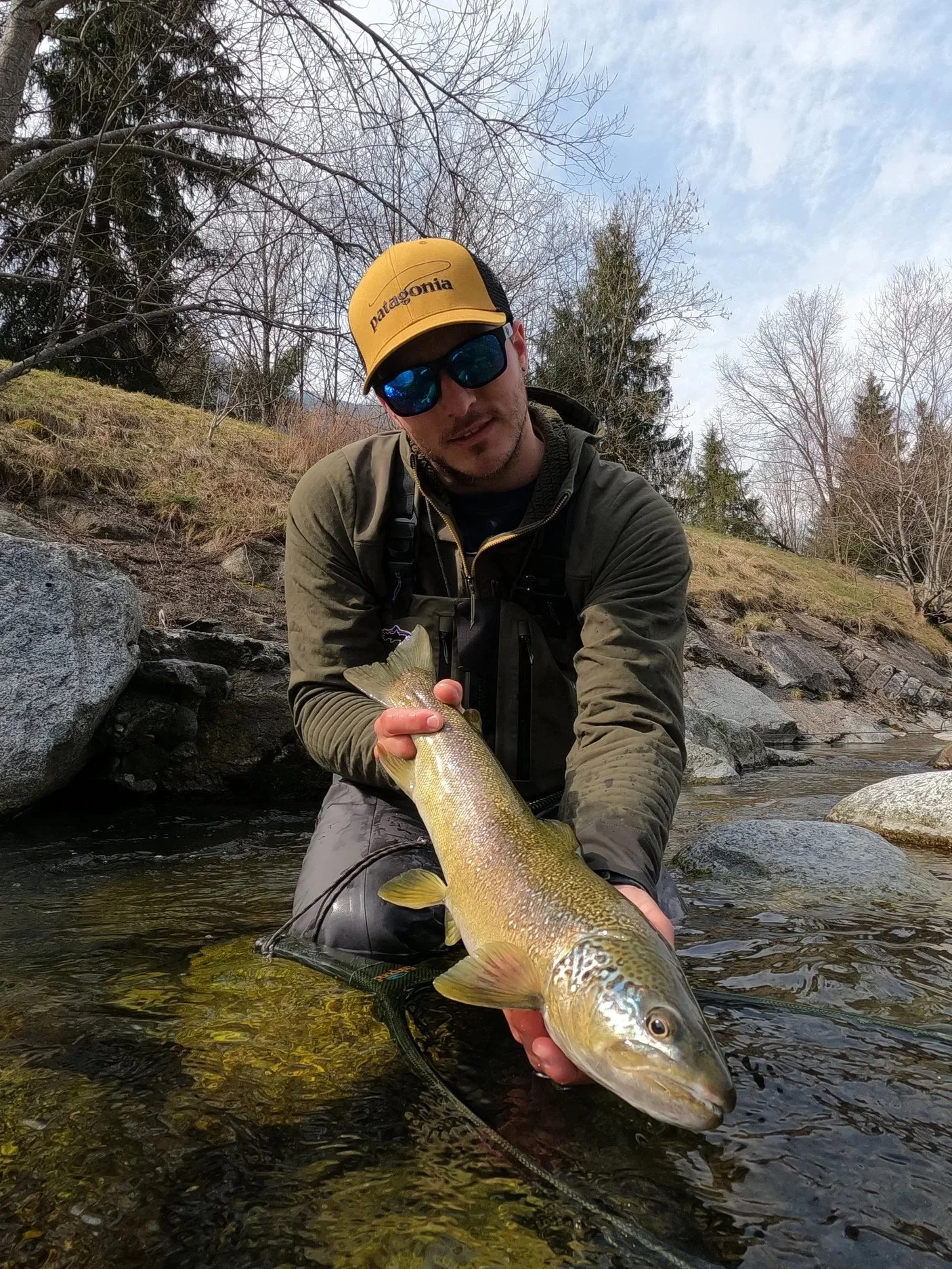 A man wearing sunglasses and a yellow Patagonia cap holds a large fish while standing in a shallow creek with rocks and leafless trees in the background.