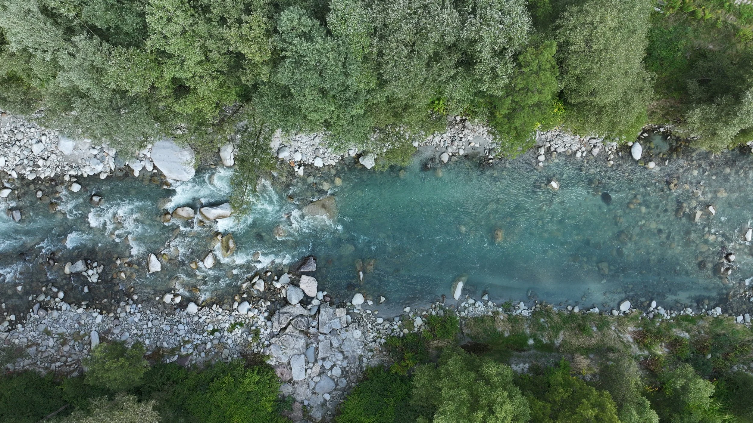 An aerial view of a river flowing through a forest with trees and rocks along the riverbanks of Sarca river.