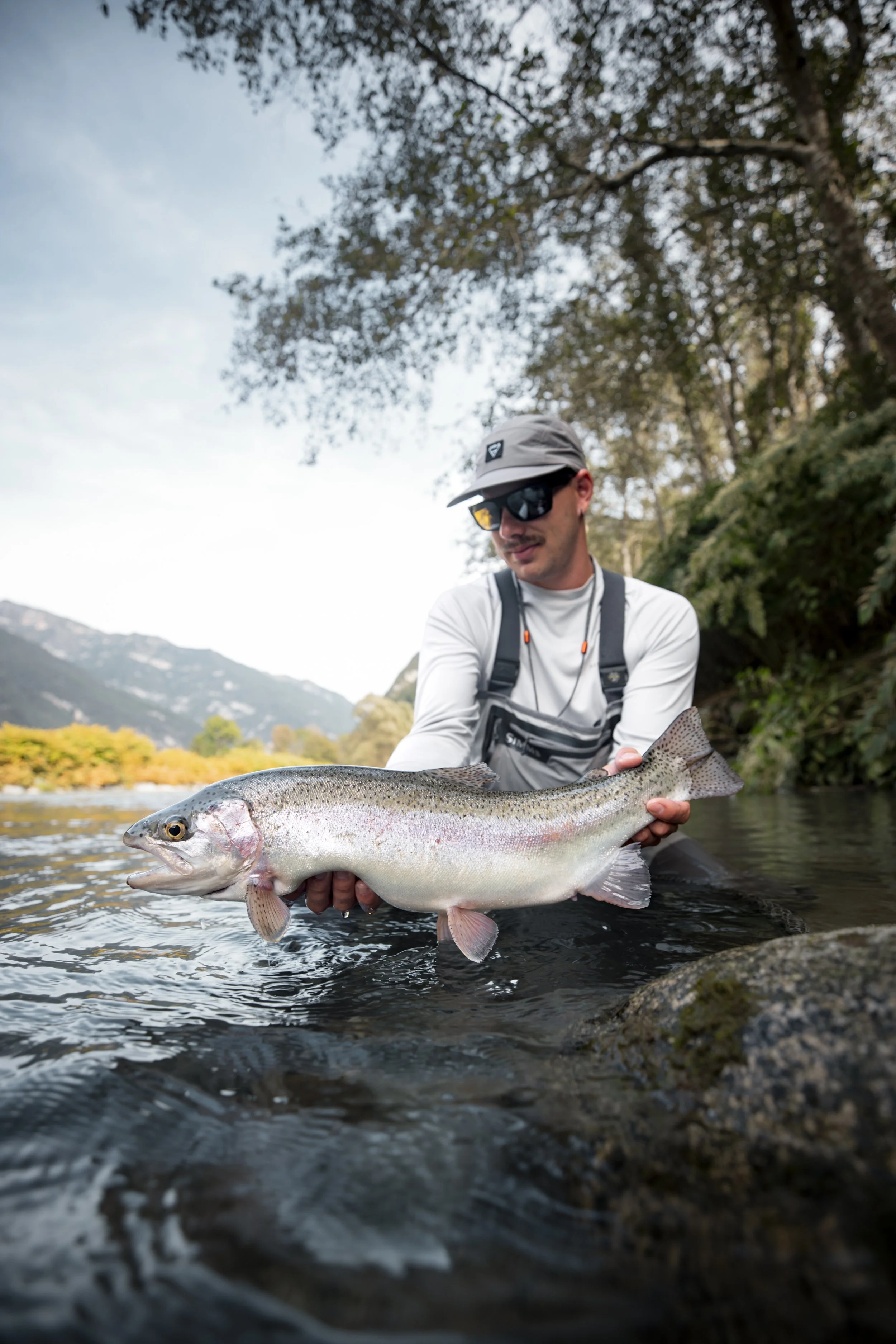 A man in sunglasses and outdoor gear holds a large rainbow trout over the water of a river, with trees and mountains in the background.