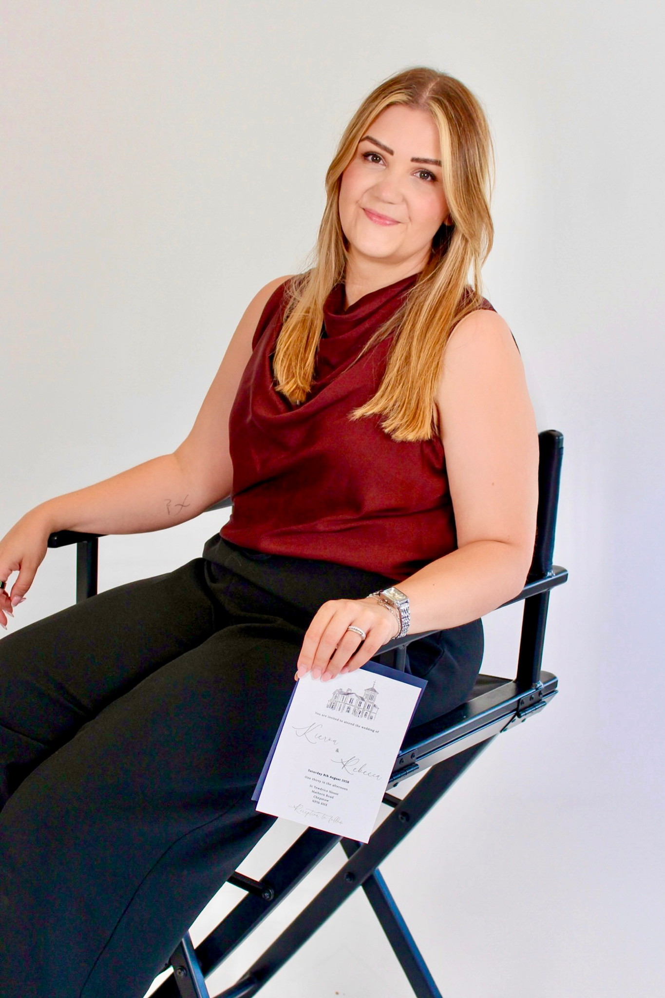 Woman with long blonde hair, wearing a sleeveless maroon top, sitting on a black chair, holding a wedding invitation, smiling at the camera.
