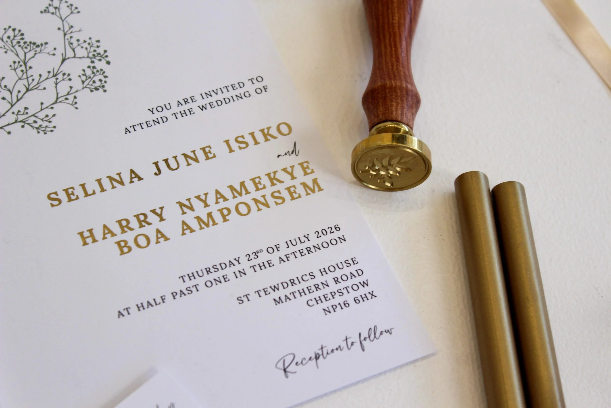 Wedding invitation with black ink text, decorated with a black botanical illustration in the top left corner, next to a wooden and gold wax seal stamp, gold-colored cylindrical object, and a wooden handle wax seal stamp.