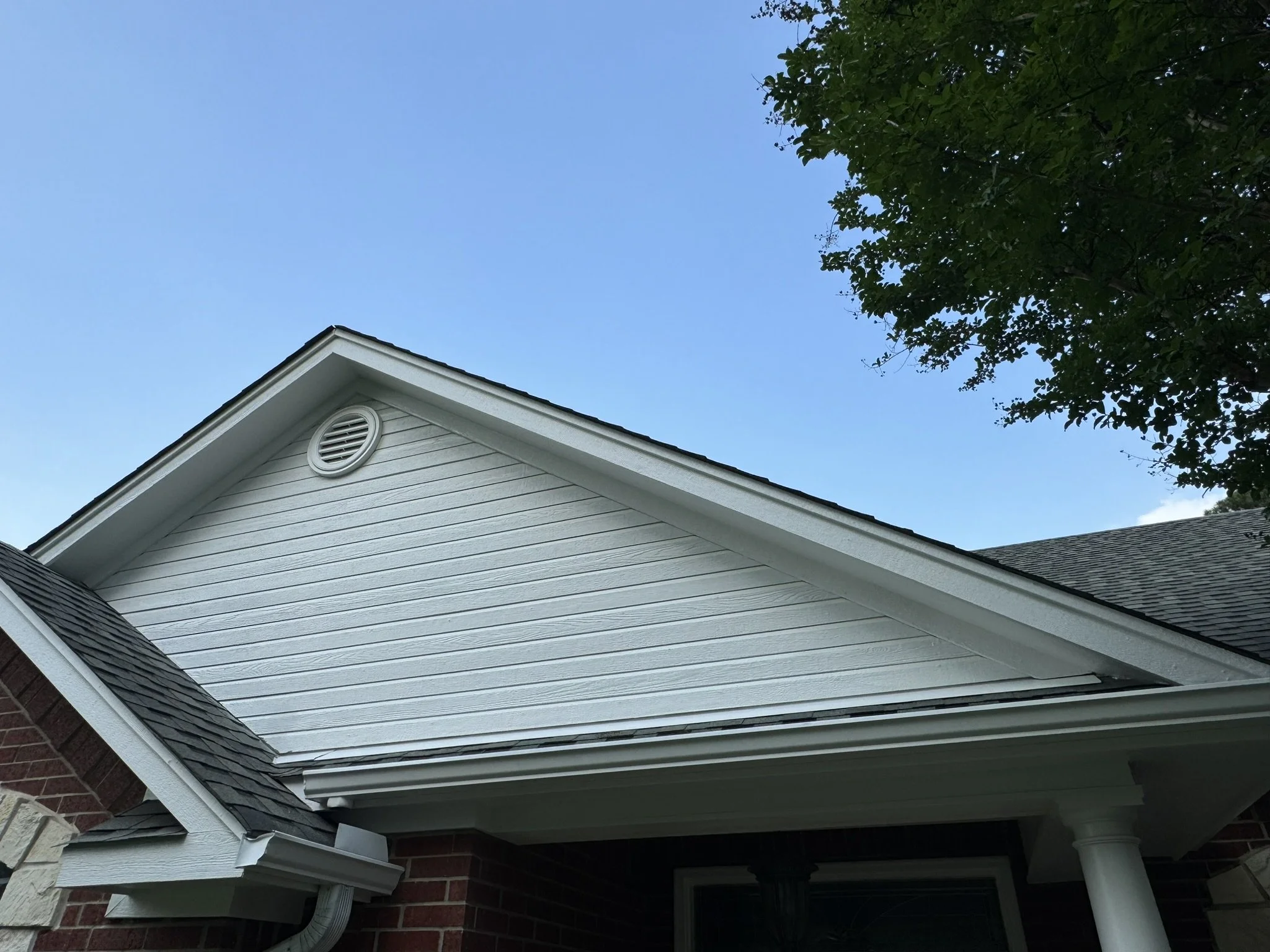 The image shows the side of a house with light gray horizontal siding, a white vent near the roof's peak, and a section of brick wall at the bottom. Part of a white gutter system is visible at the lower left corner. The sky above is clear and blue, a