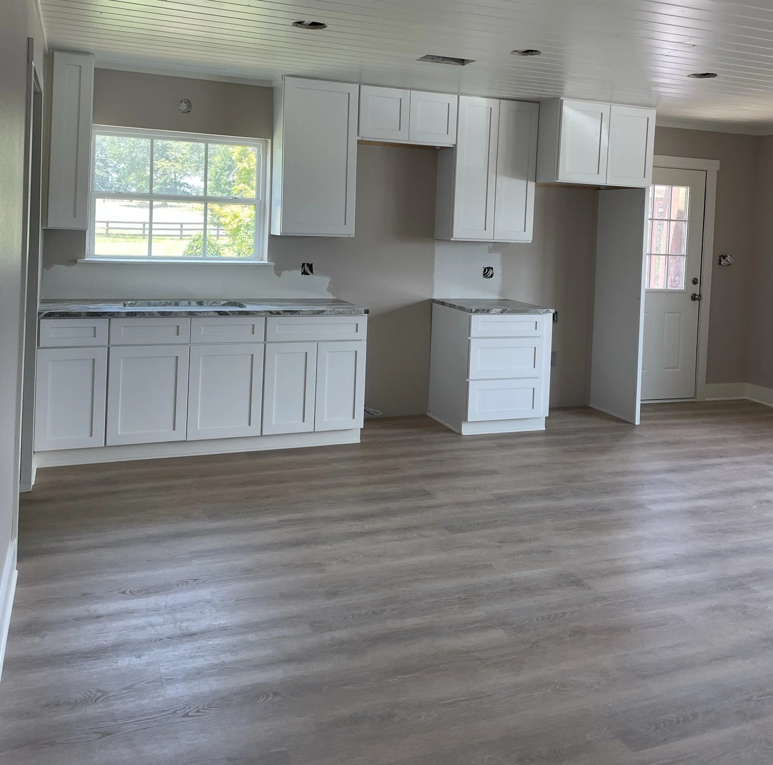 Empty kitchen with white cabinets, a window overlooking greenery, and light wood flooring.