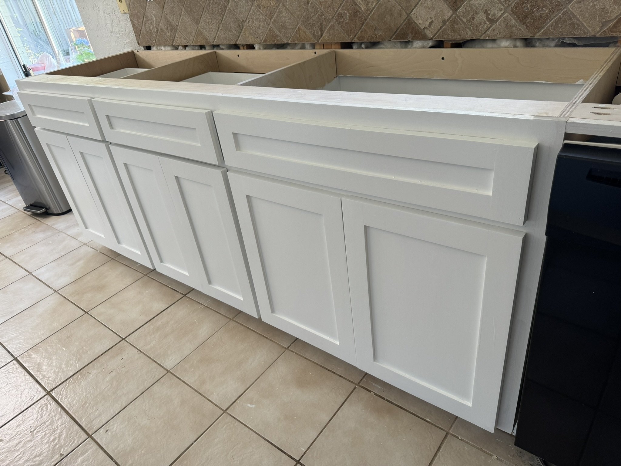 Kitchen cabinet with white doors, incomplete countertop with exposed wood and cabinet framing, tile floor, and a window on the left side.