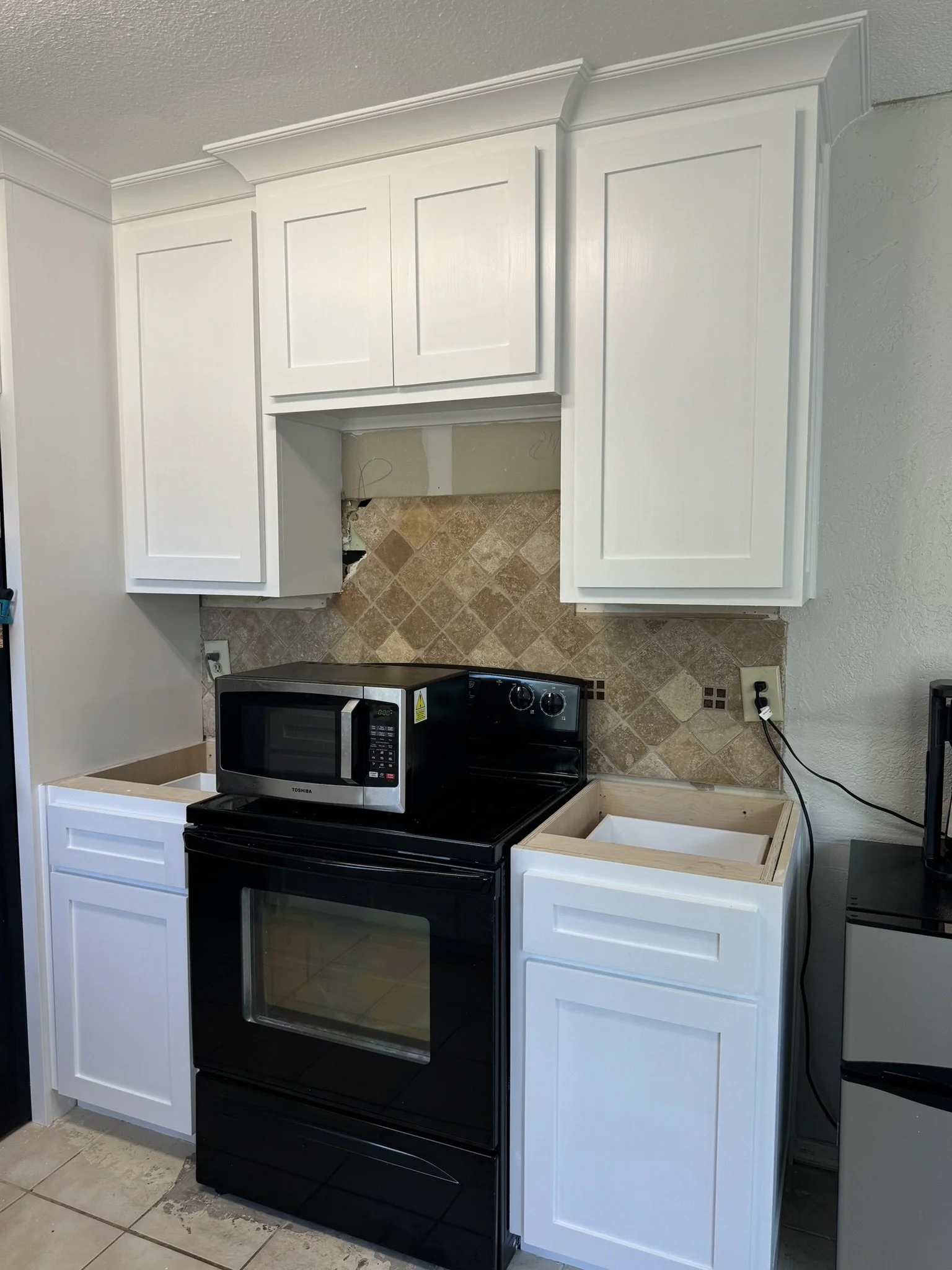 Kitchen with white cabinets, black stove and microwave, and beige tiled backsplash under construction.