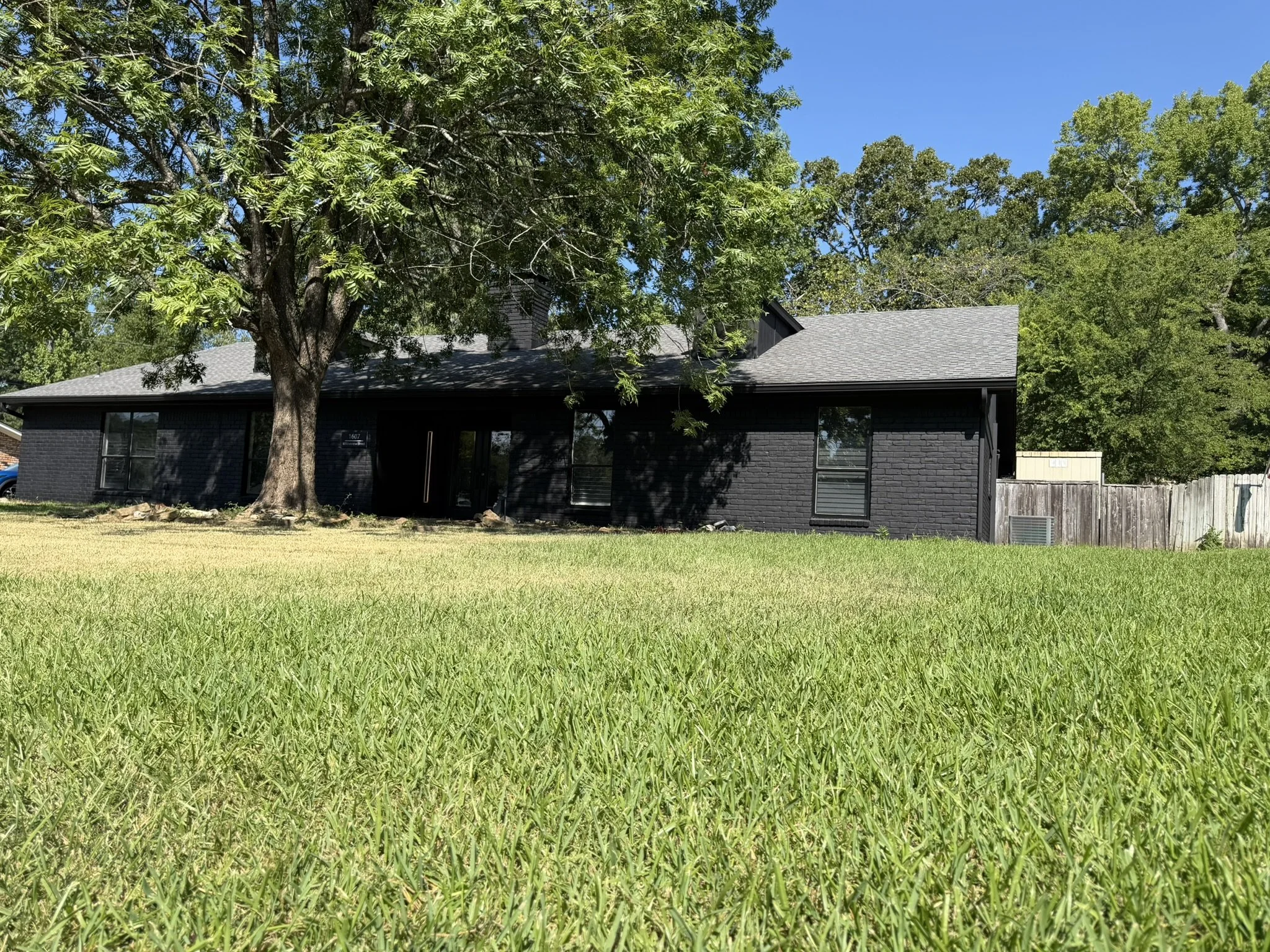 A black house with a gray roof, large trees in front, and a grassy lawn with a clear blue sky overhead.