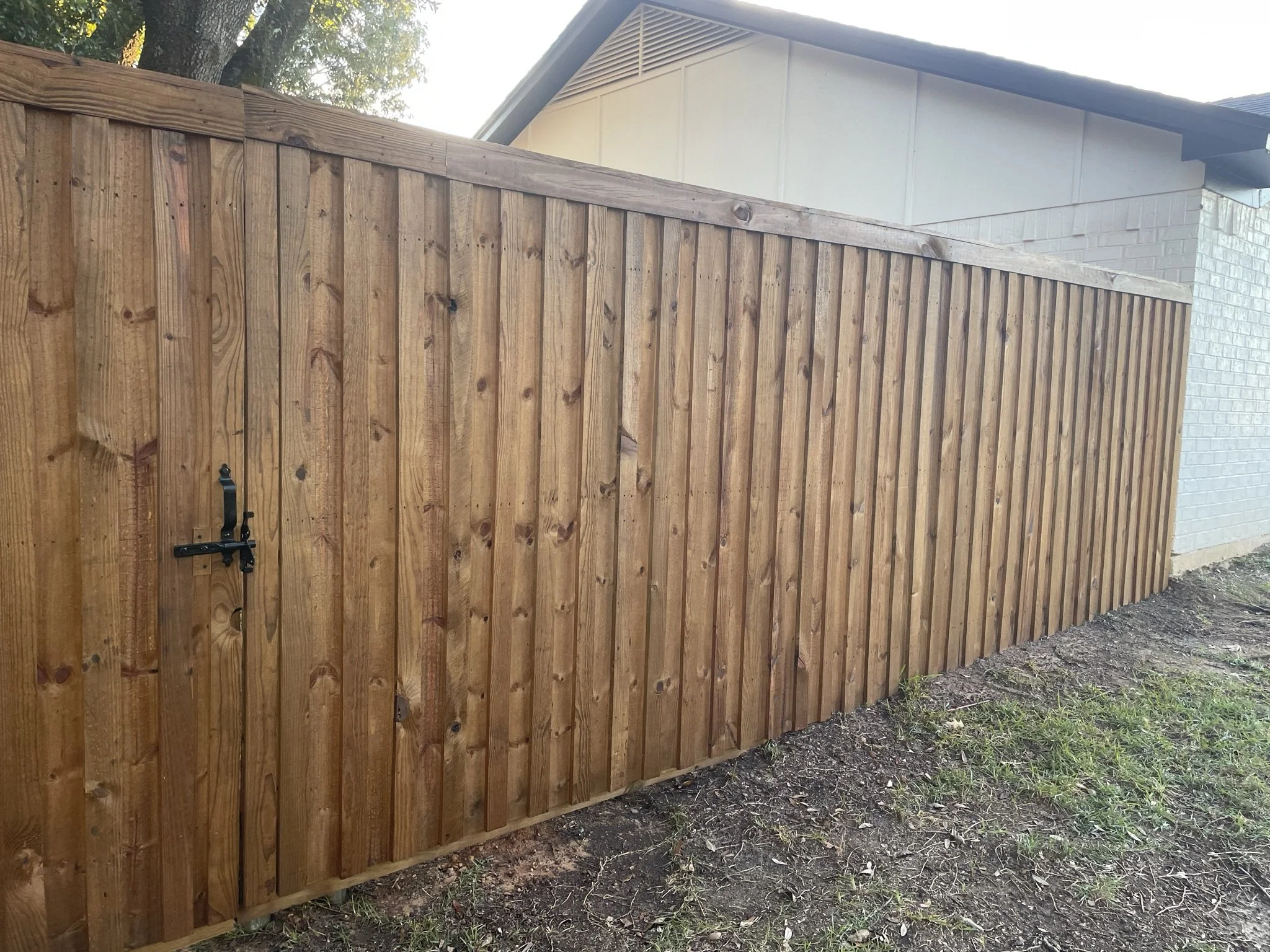 A newly installed wooden privacy fence with vertical planks and a black latch gate, next to a house with partial white brick and siding exterior, and a tree in the background.