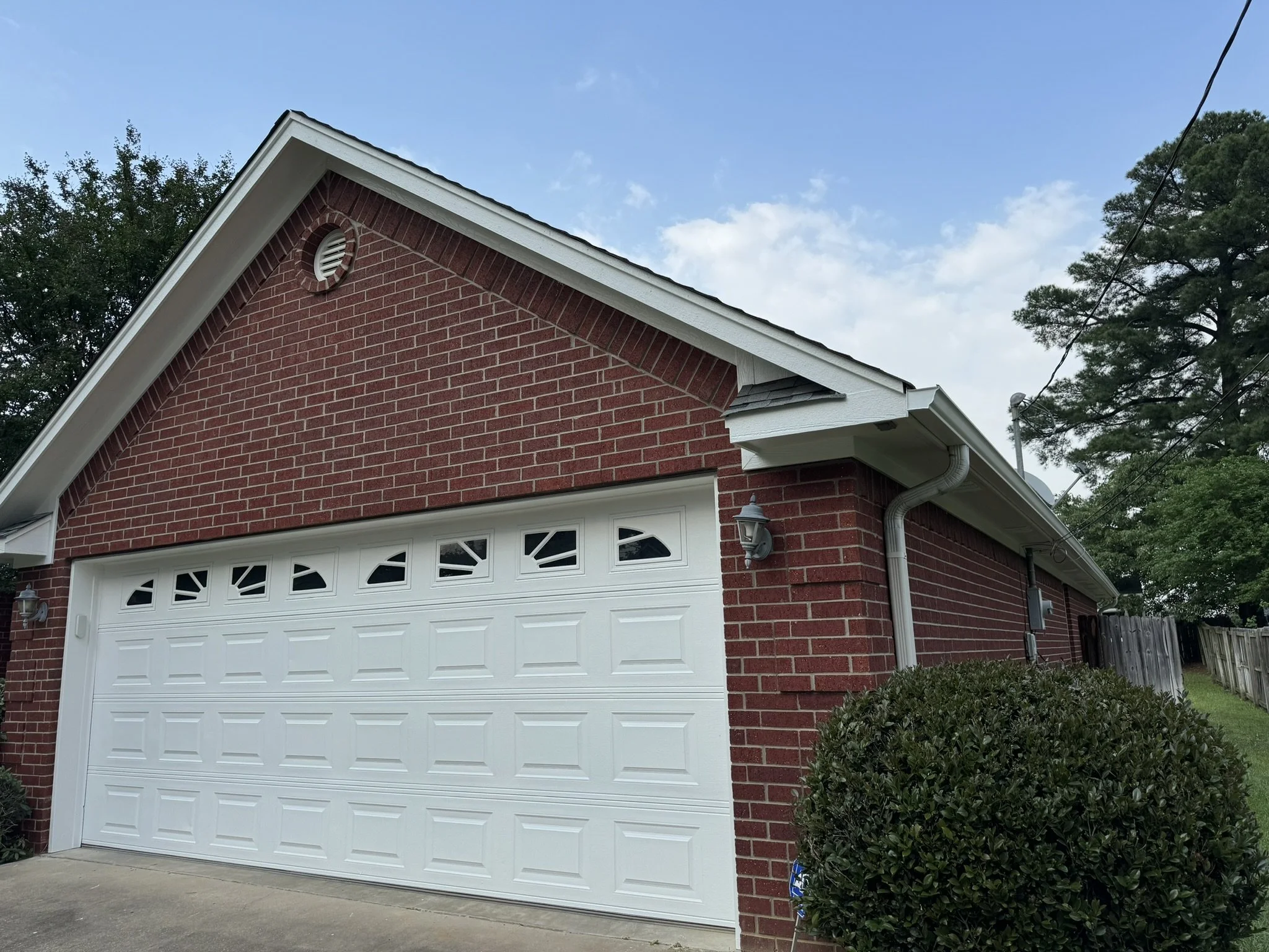 Red brick house with white garage door, black shingled roof, two outdoor lantern lights, bush in front, large trees in background, clear blue sky with some clouds.