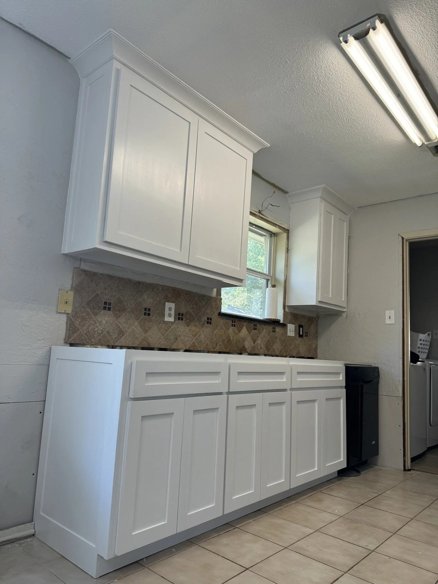 White kitchen cabinets with a beige tiled backsplash, a window above the countertop, and a fluorescent light on the ceiling.