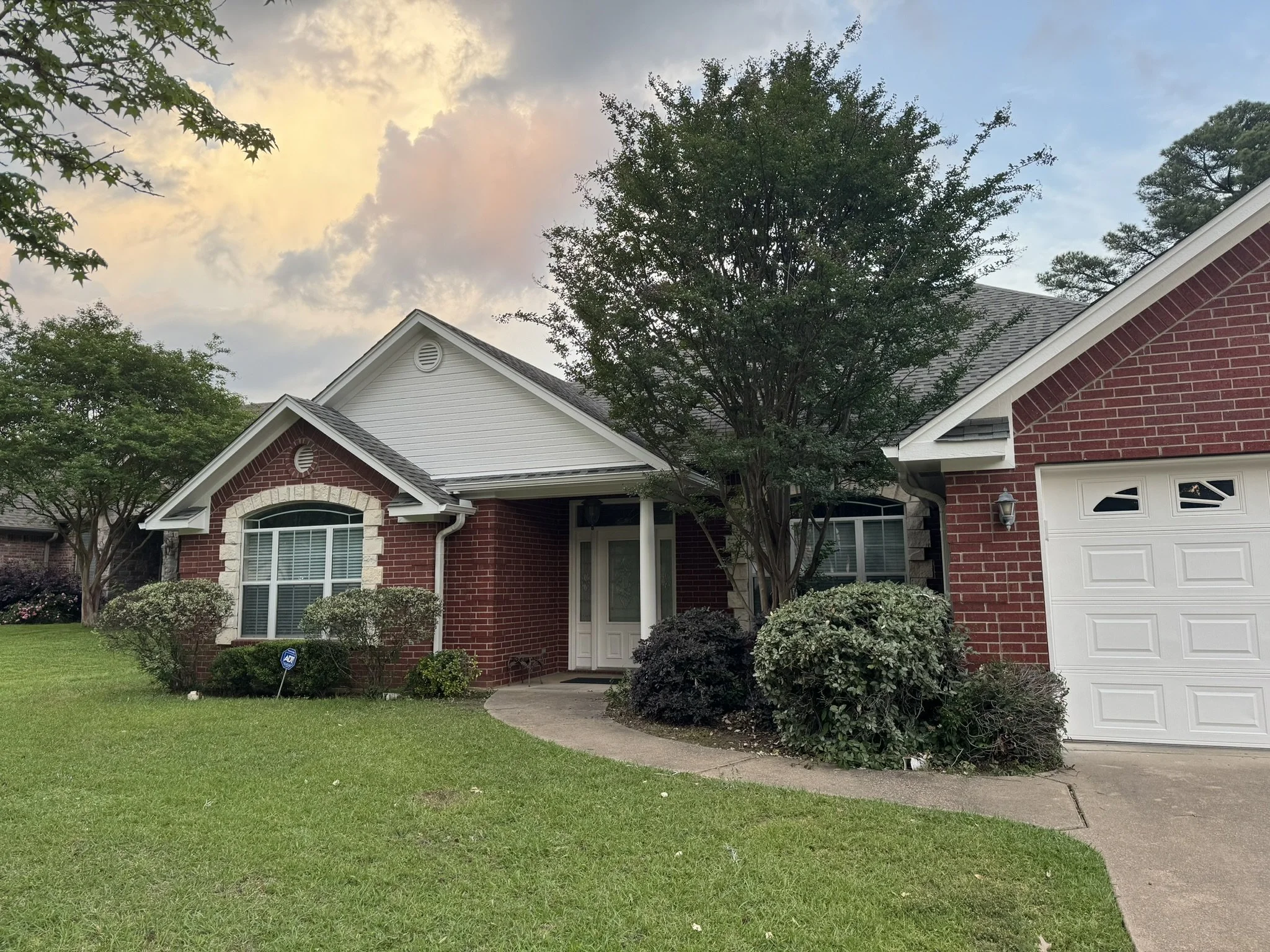 Front view of a brick house with a white garage door, surrounded by green trees and shrubs, under a partly cloudy sky at sunset.