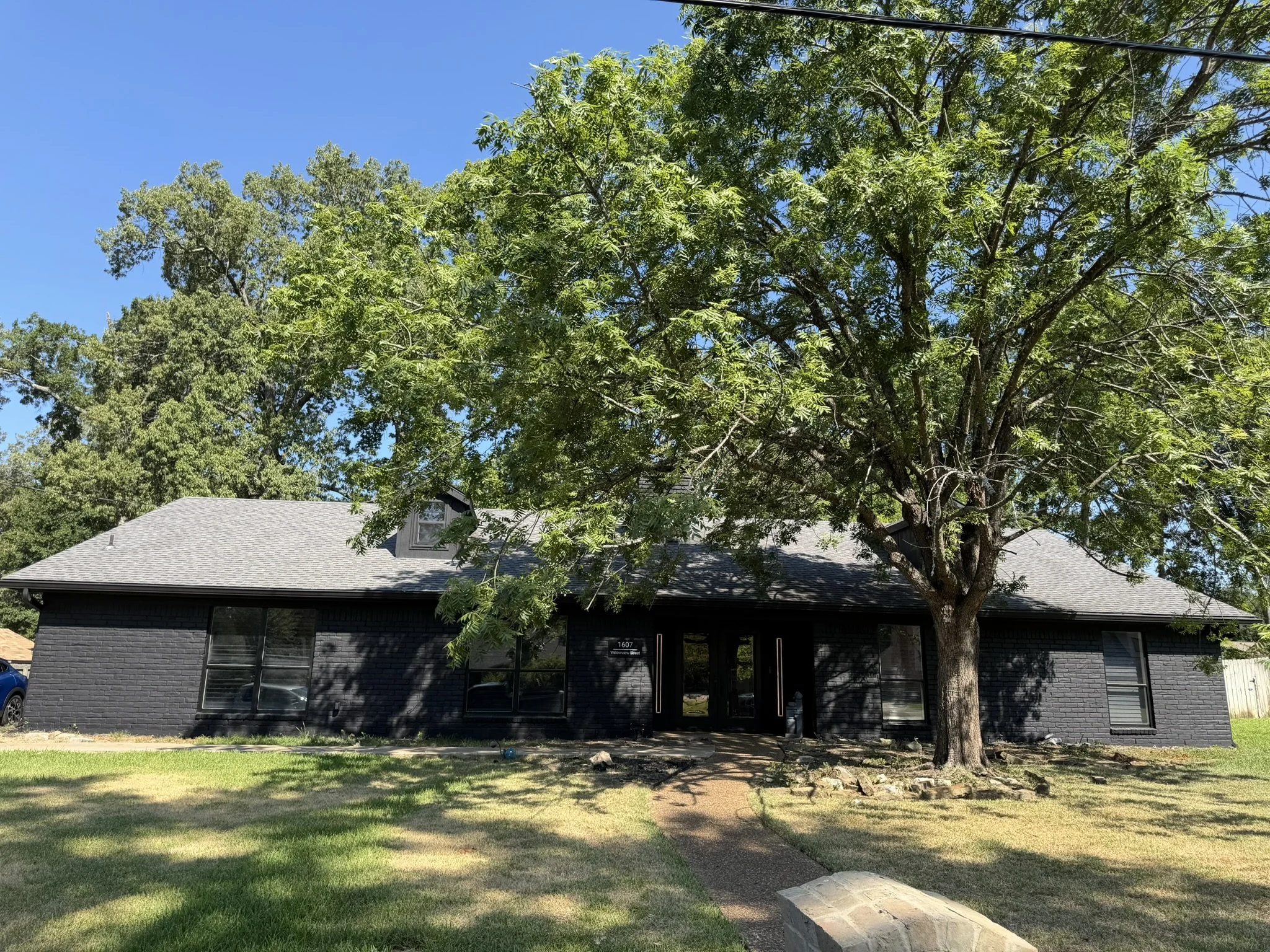 Modern black brick house with large windows, surrounded by a green lawn and a tall leafy tree in front under a clear blue sky.