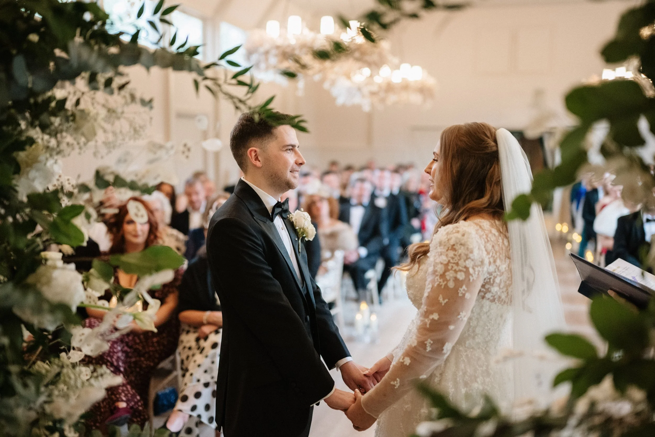 A bride and groom holding hands during their wedding ceremony, facing each other in a decorated indoor venue with guests watching.