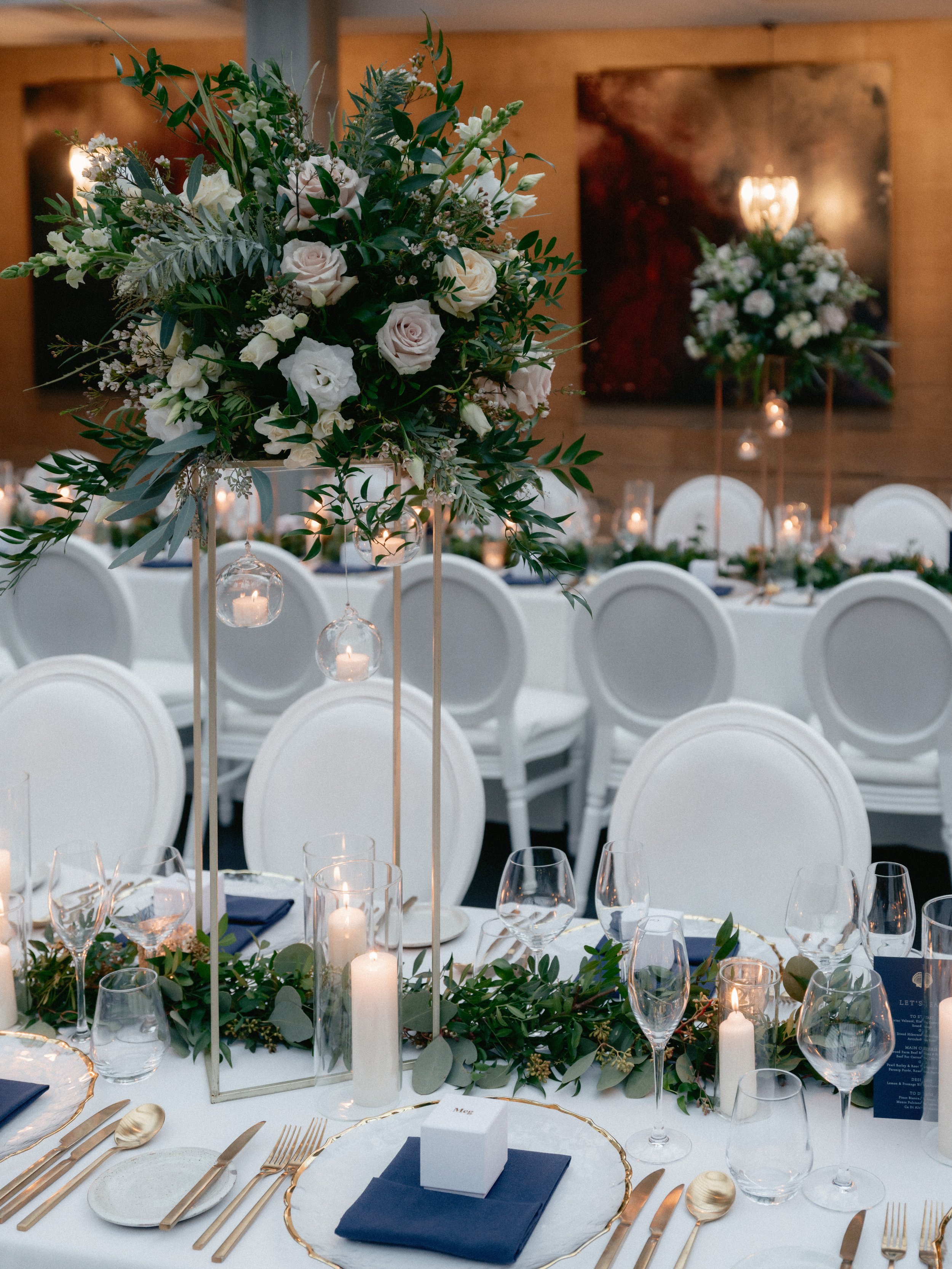 Elegant dining table set with white linens, candles, glassware, floral arrangements, and brushed gold cutlery. 