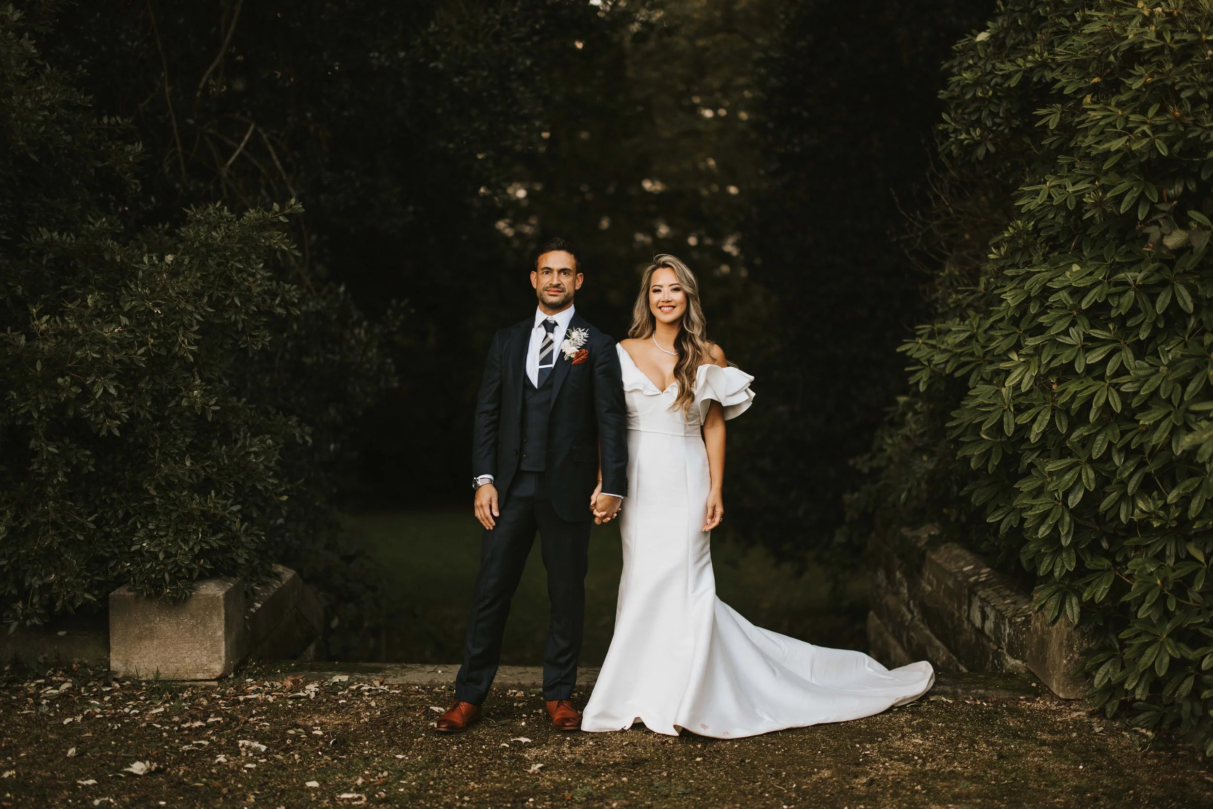 A bride and groom stand hand in hand outdoors, surrounded by lush greenery, on their wedding day. The bride wears a white gown with ruffled sleeves, and the groom wears a dark suit with a white shirt and striped tie.