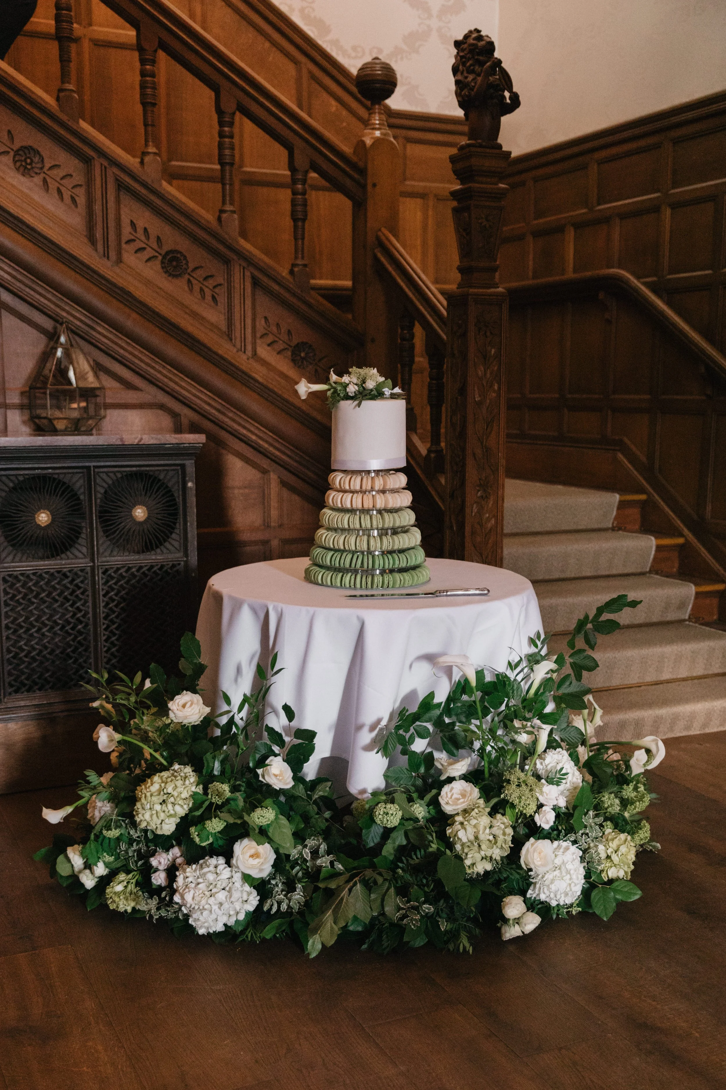 Elegant wedding cake on a round table, decorated with green and white macarons and topped with flowers, surrounded by white and green floral arrangements, in a wood-paneled hall.