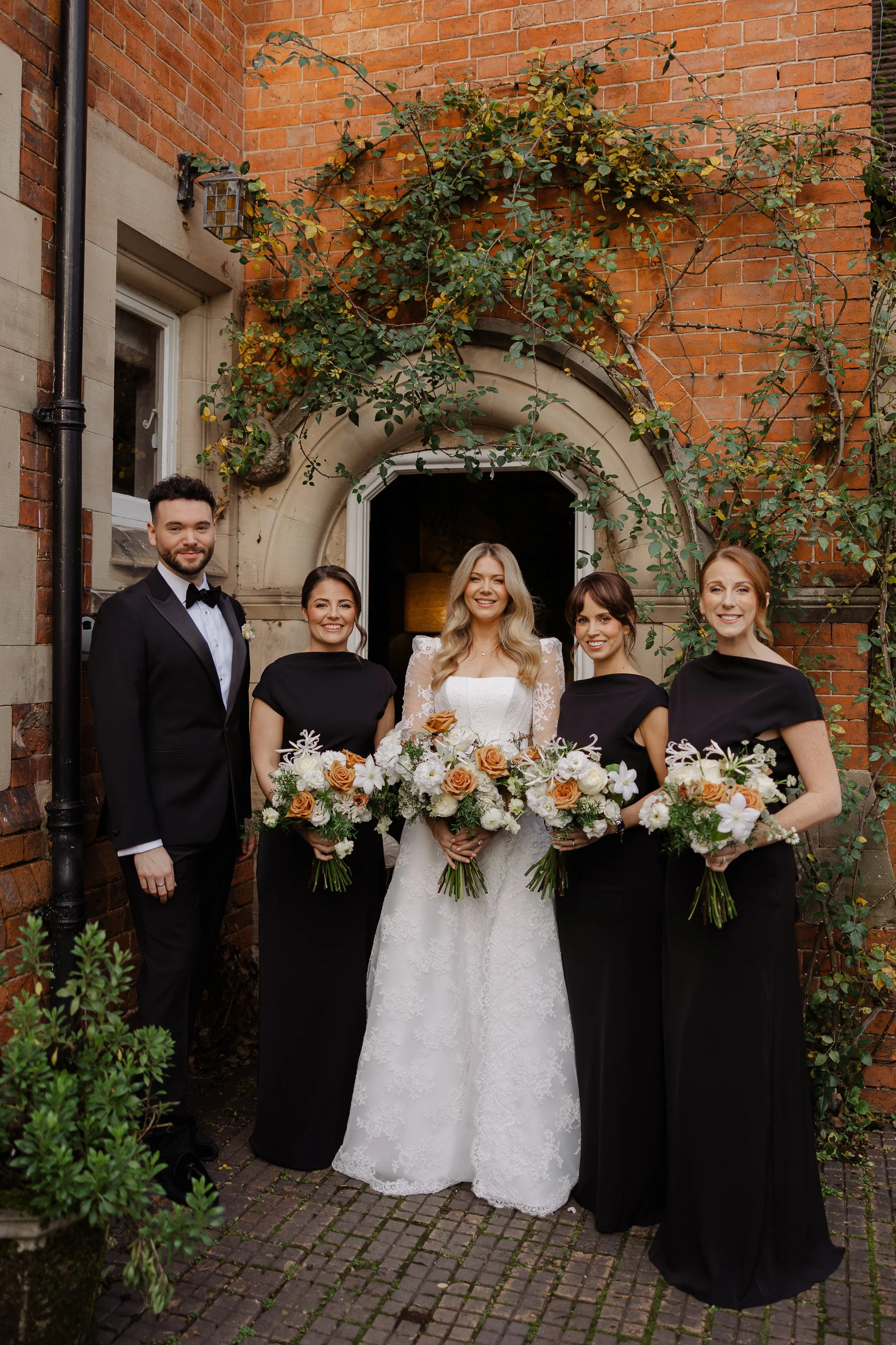 A bride standing with four bridesmaids and a groomsman outside a brick building with green vines, holding bouquets of white and beige flowers.