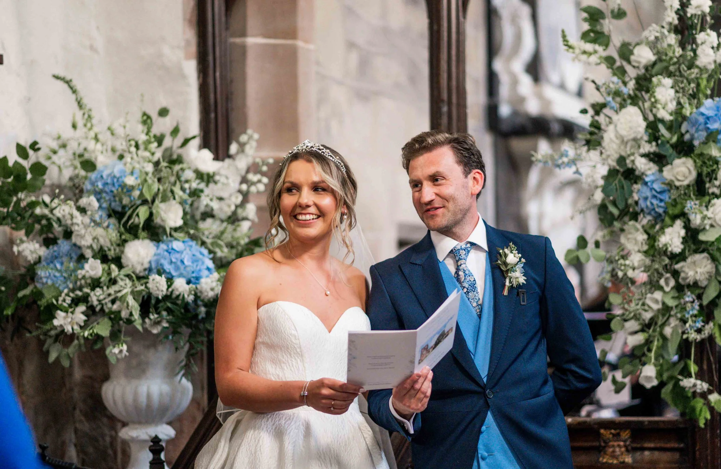 A bride and groom at their wedding ceremony, standing in front of large floral arrangements of white and blue flowers, inside a church or historic building.
