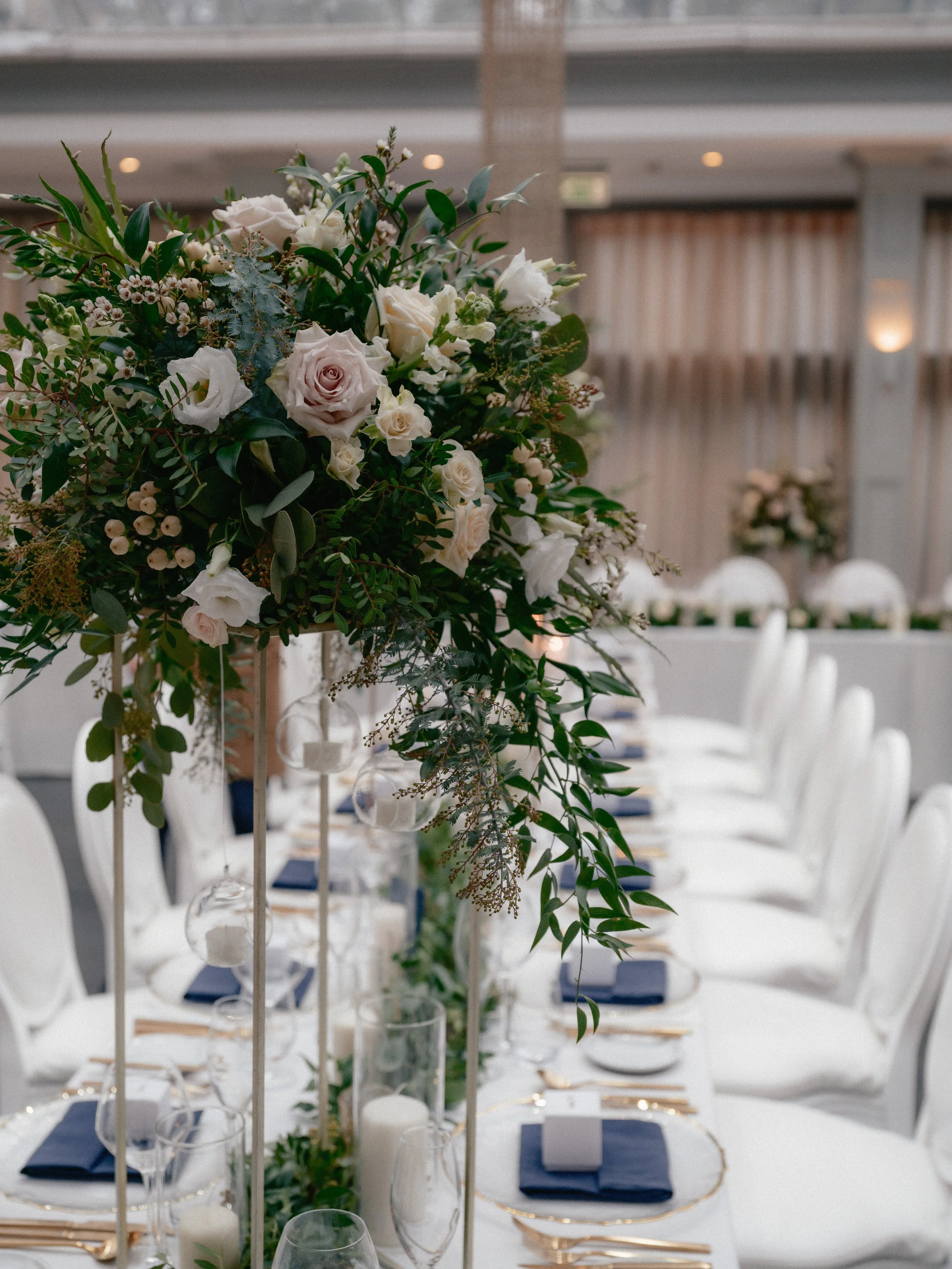 Elegant banquet table decorated with a tall floral centerpiece of white and pastel roses, greenery, candles, and table settings with white plates, navy blue napkins, and gold flatware.