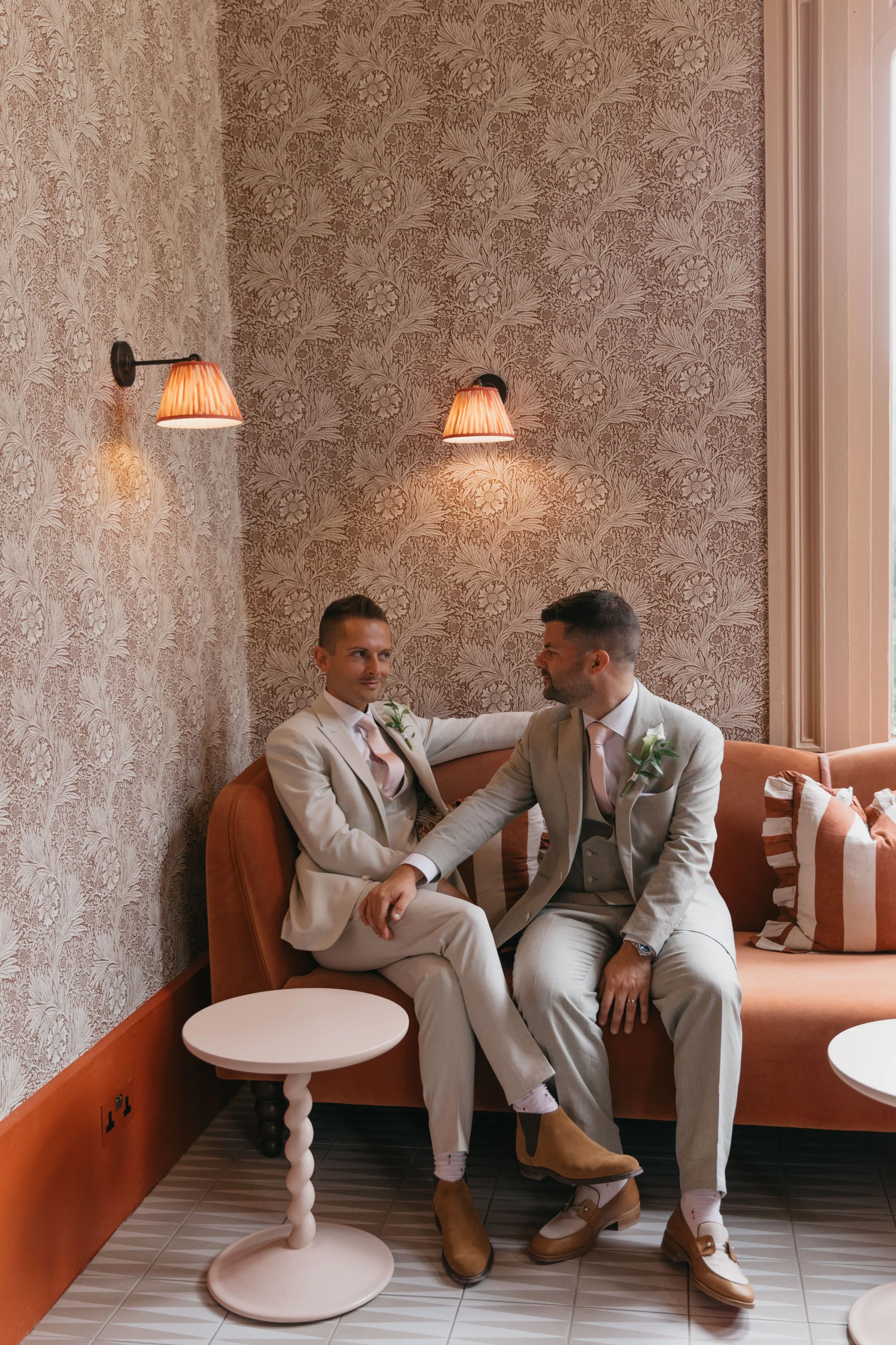 Two grooms in beige suits sitting on a pink couch in a vintage-style room with floral wallpaper, facing each other. One man has his arm around the other, and they are holding hands, engaging in an intimate moment.