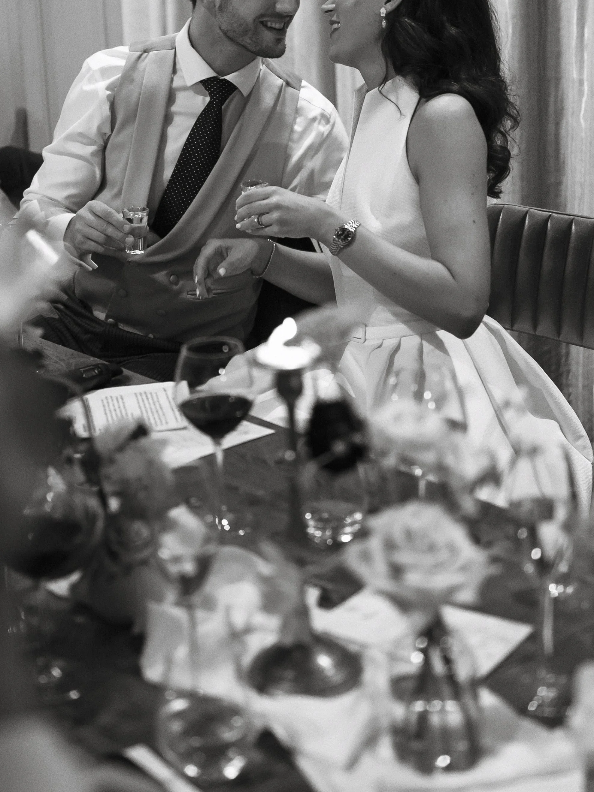 A smiling couple at a dinner table, holding shot glasses, dressed in formal attire, with wine glasses and floral centerpieces in the foreground.