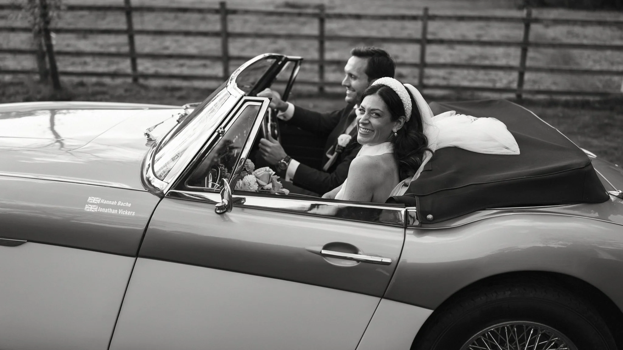 Bride and groom smiling and riding in a vintage convertible car, with a wooden fence in the background.